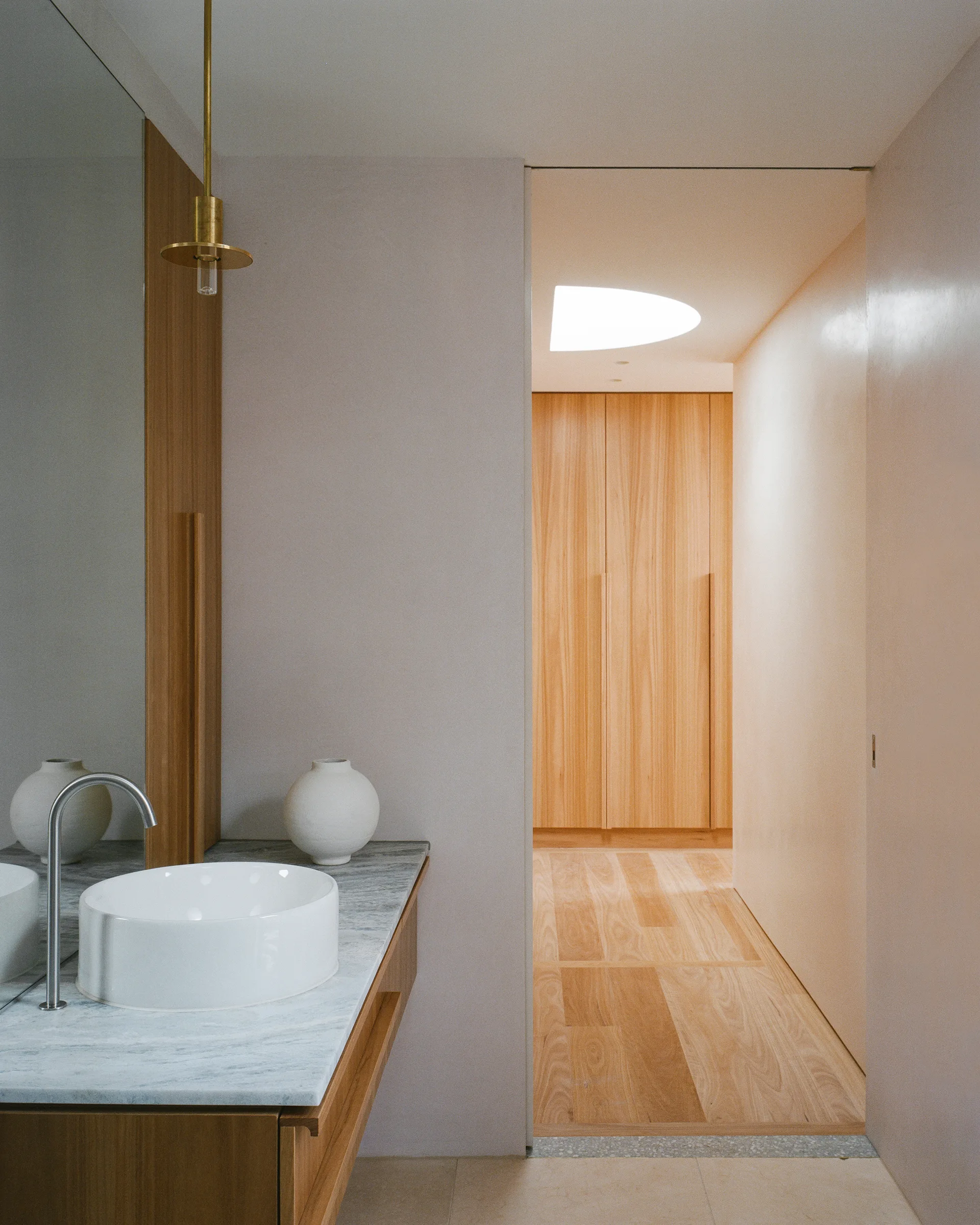 A minimalist bathroom vanity with a white vessel sink and marble countertop near a hallway with natural wood floors and built-in wood closets.