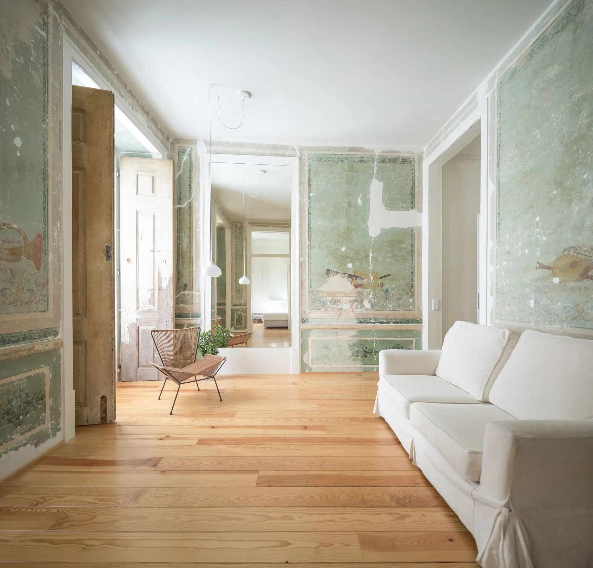 A minimalist living room with distressed sage green fresco walls, a white slipcovered sofa, a modern woven lounge chair, and light wood flooring.