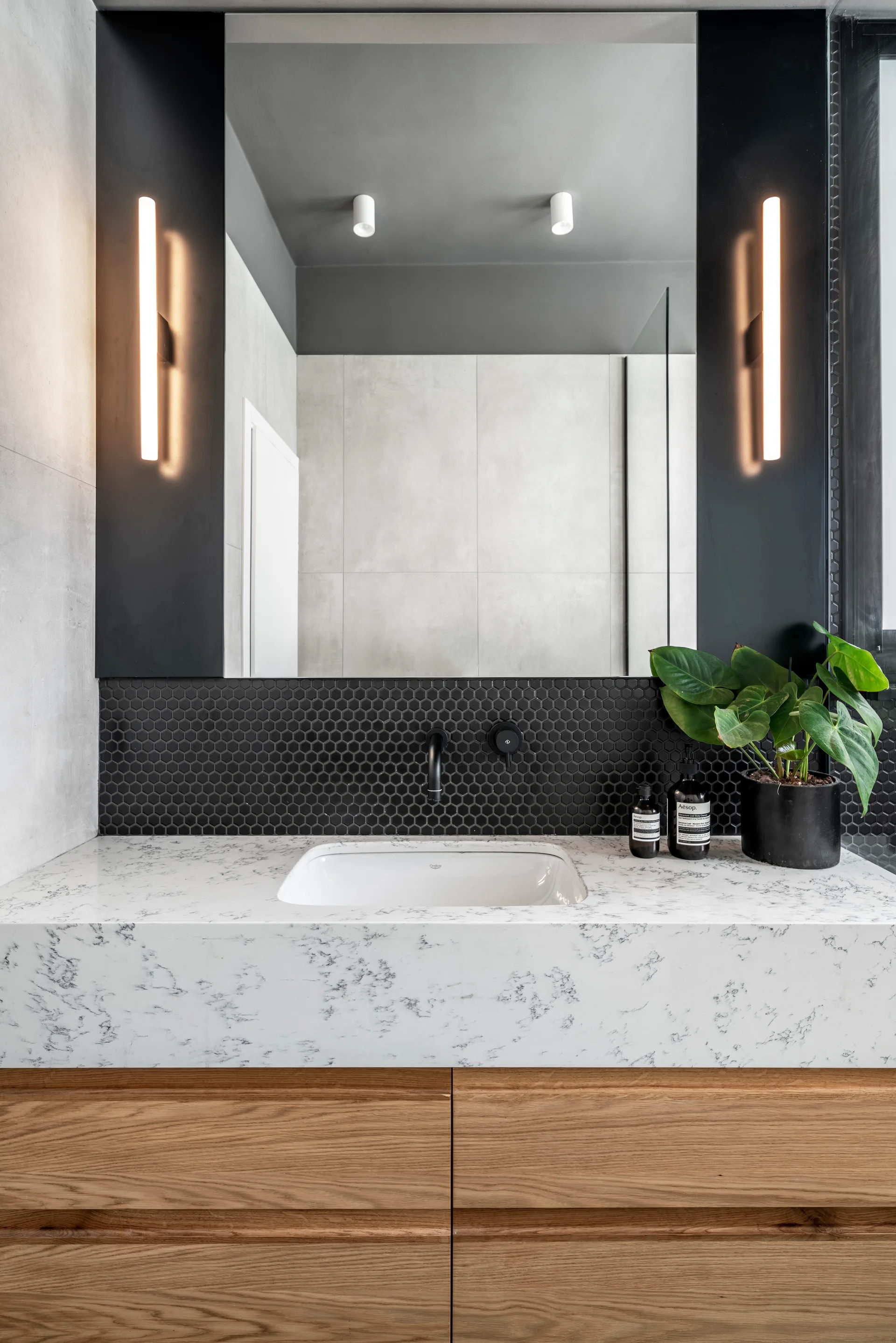 A modern bathroom vanity features a wood cabinet, white marble countertop, a black hexagonal tile backsplash, and a wall-mounted black faucet.