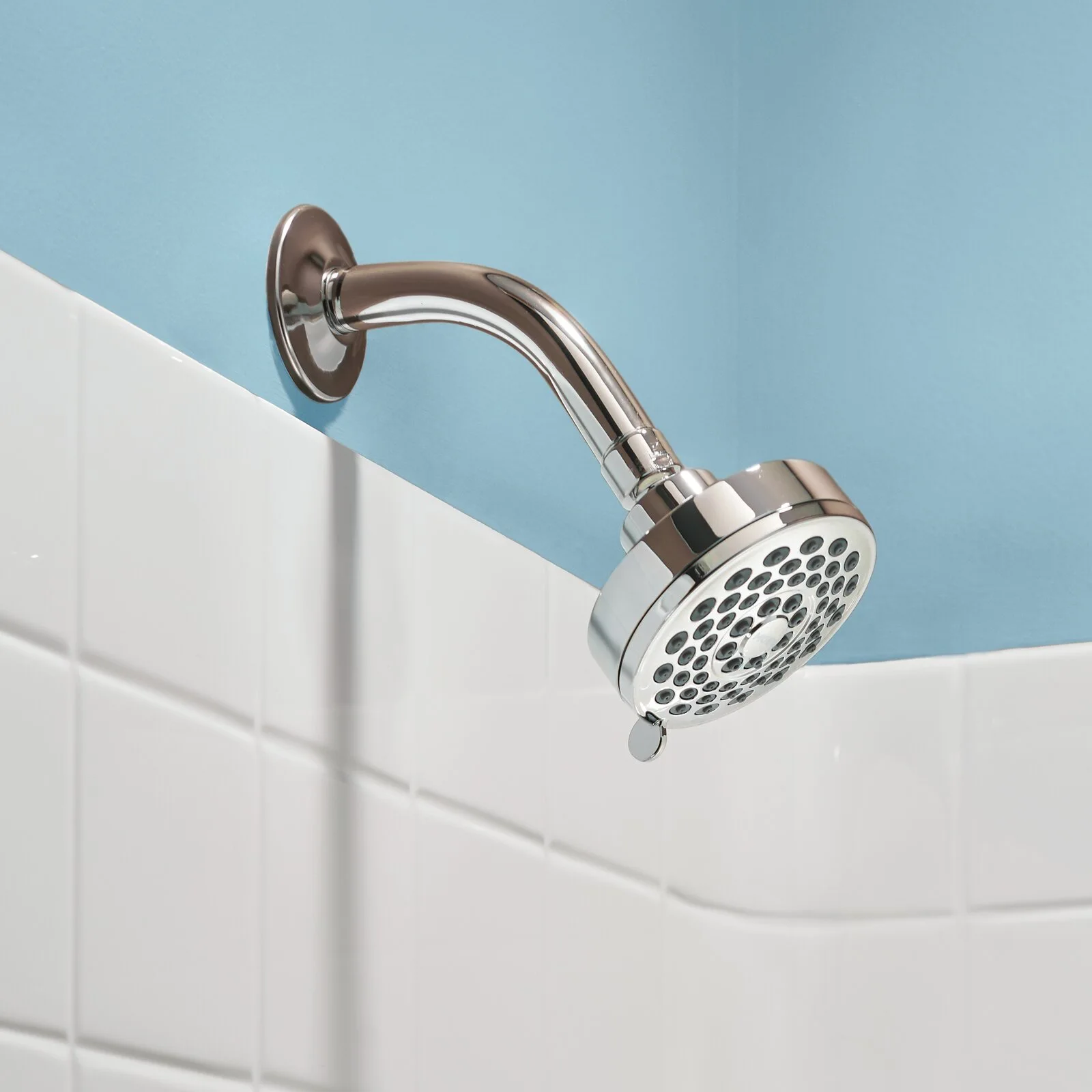 A polished chrome shower head is mounted on a wall covered with white square tiles below a light blue painted surface.