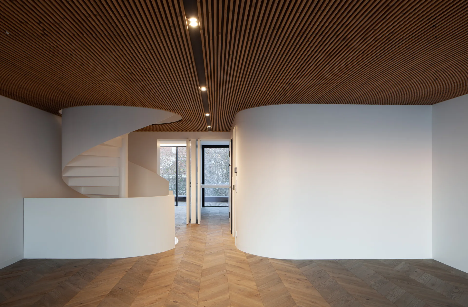 A minimalist hallway features a white spiral staircase, curved white walls, a warm slatted wood ceiling, and herringbone pattern hardwood flooring.