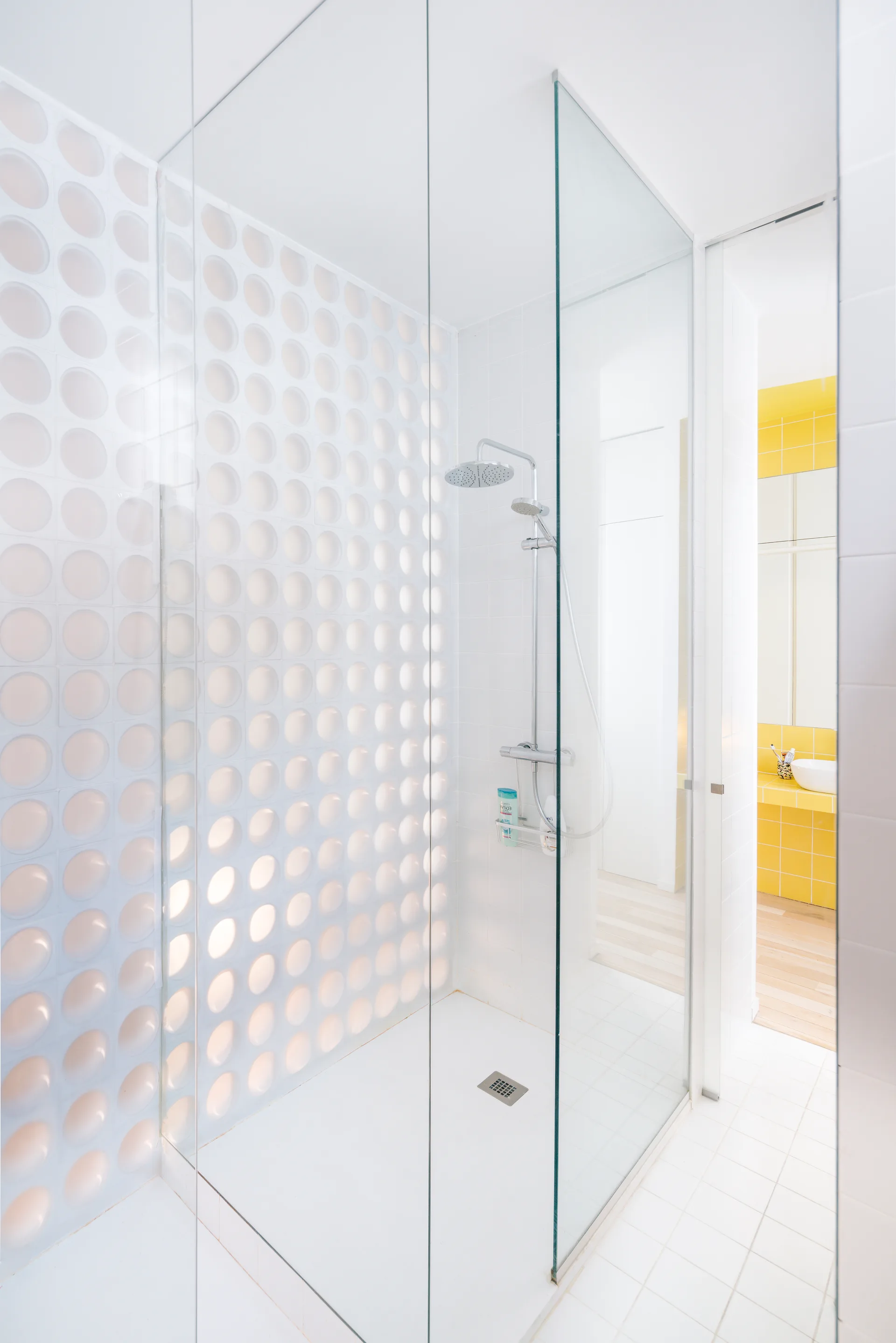 A modern walk-in shower with clear glass panels, unique circular-relief white wall tiles, and a view of a yellow tiled bathroom counter.