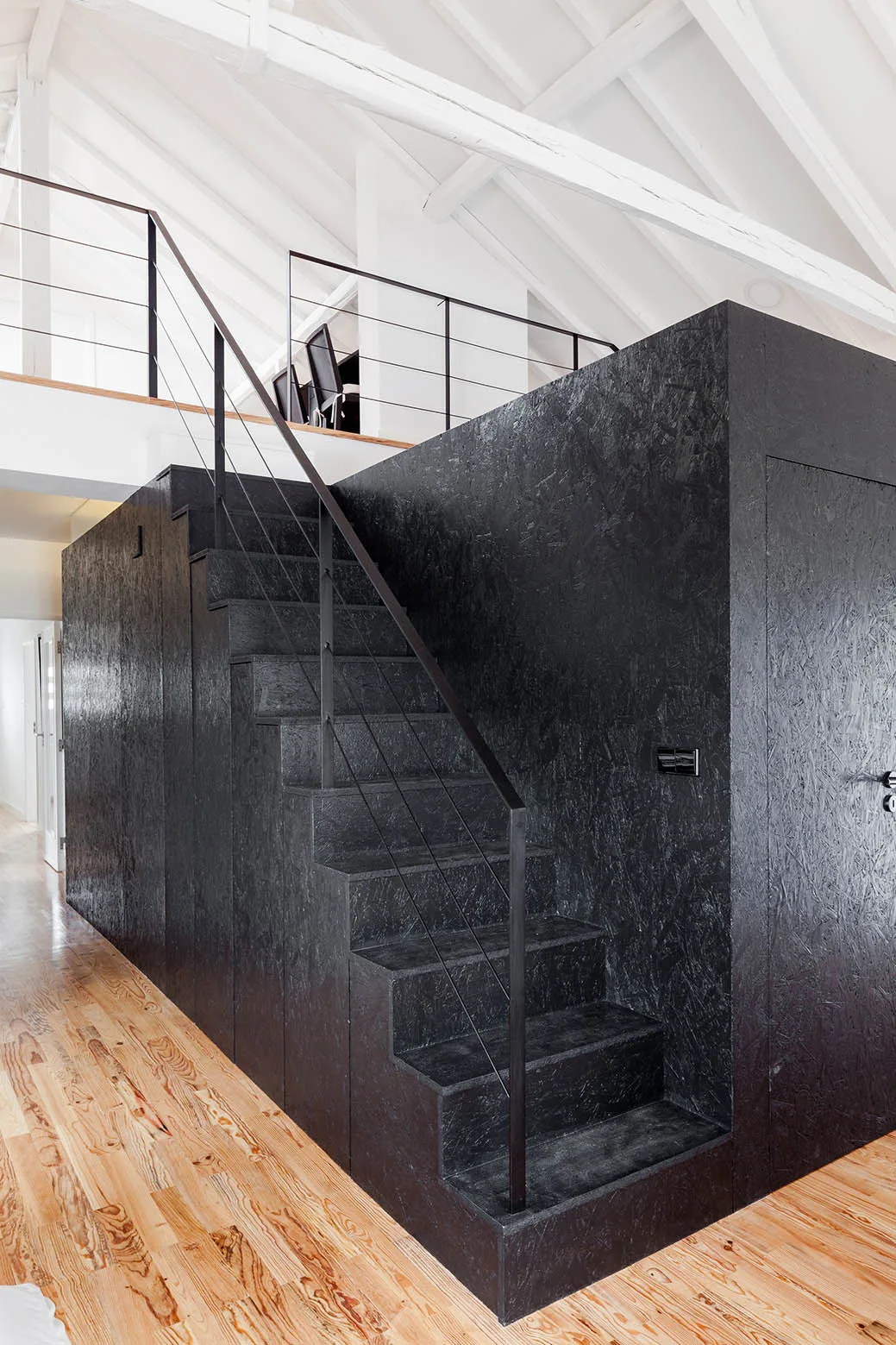 A black oriented strand board staircase with a wire railing rises from light wood floors to a mezzanine under white painted ceiling beams.