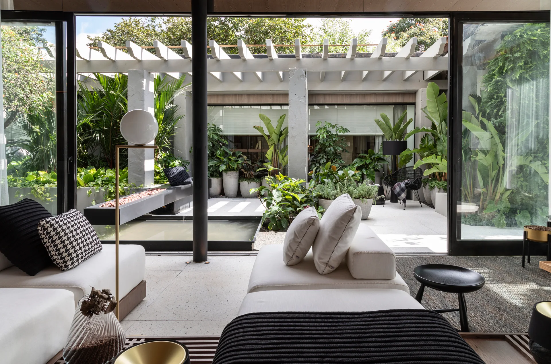 A modern living room with white modular sofas and large sliding glass doors opening onto a lush courtyard with a water feature and pergola.