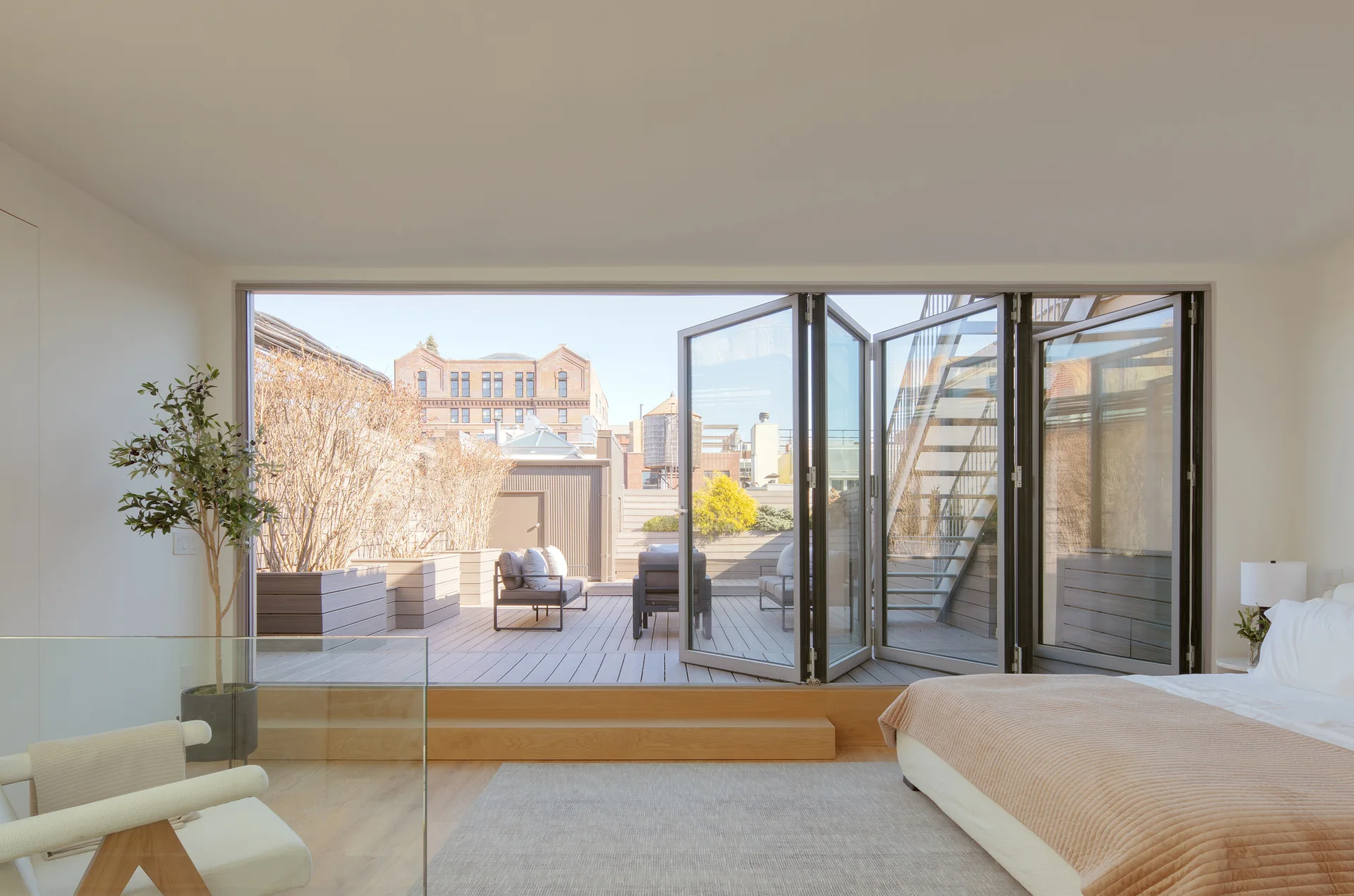 A minimalist bedroom features a bed with beige linens and large folding glass doors that open onto a wood-planked rooftop terrace.
