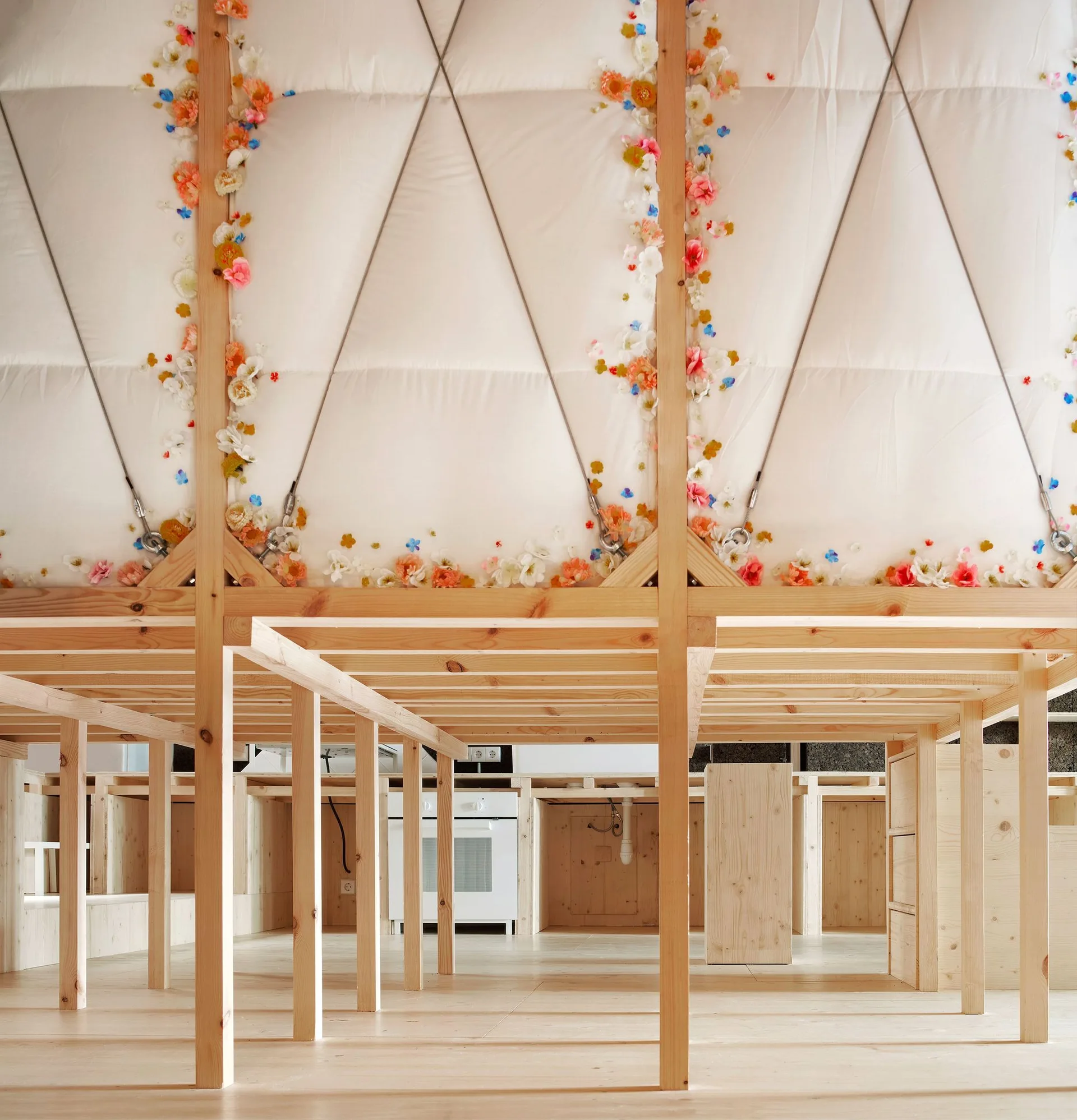 An interior view of an open wooden post-and-beam structure on light wood floors, topped by white triangular fabric panels decorated with small colorful flowers.