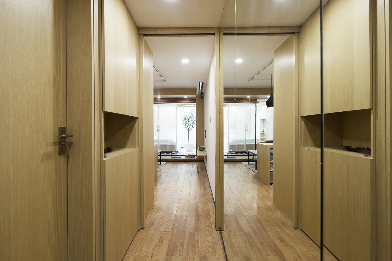 A narrow hallway features light wood paneled cabinets, wood plank flooring, and a large mirrored wall reflecting the passage and adjacent rooms.