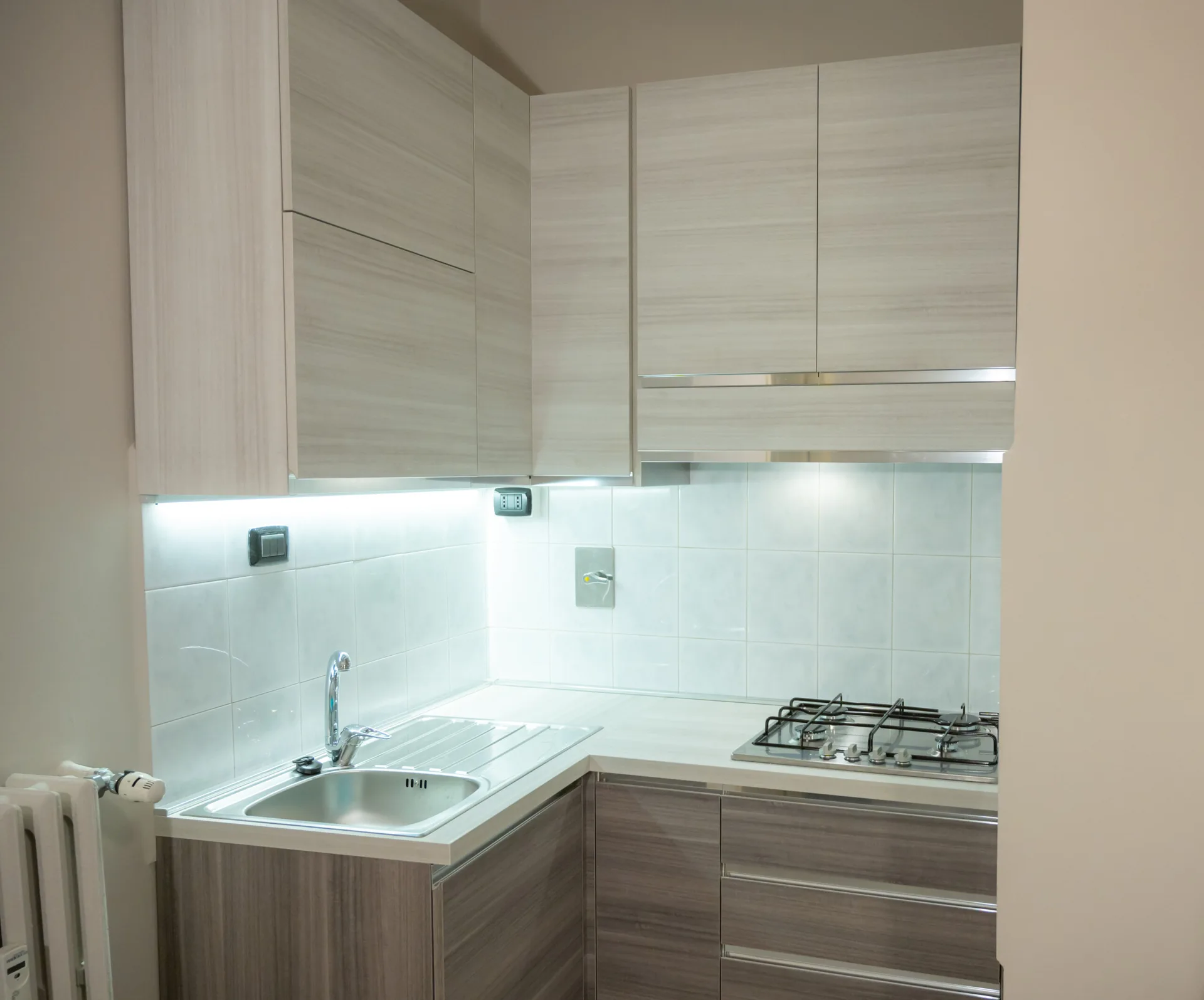 A small kitchenette features two-tone wood-grain cabinets, a white square-tile backsplash, and integrated stainless steel sink and gas stovetop.
