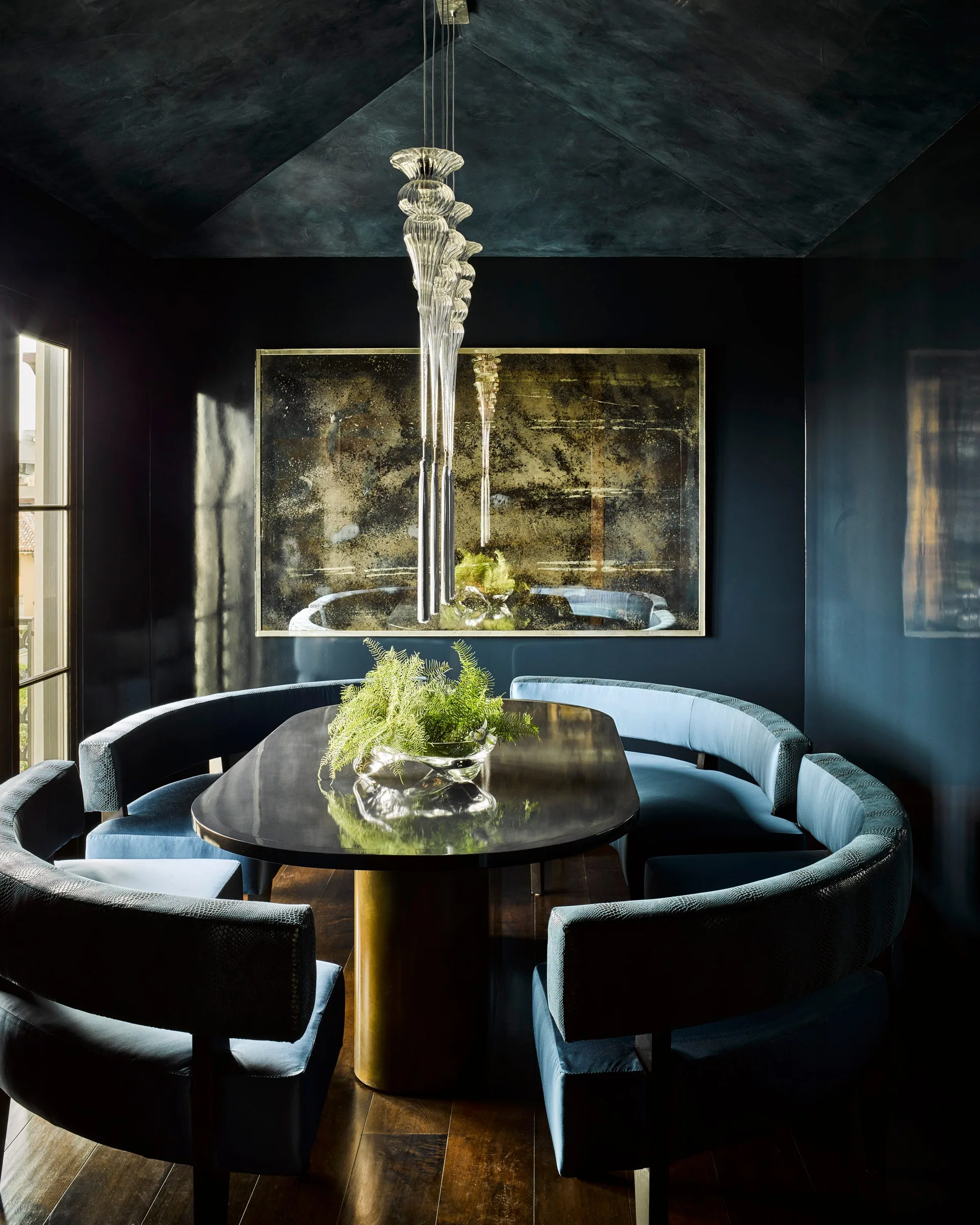 A dark navy dining room with a curved blue velvet banquette around a pedestal table beneath a tiered glass pendant light.