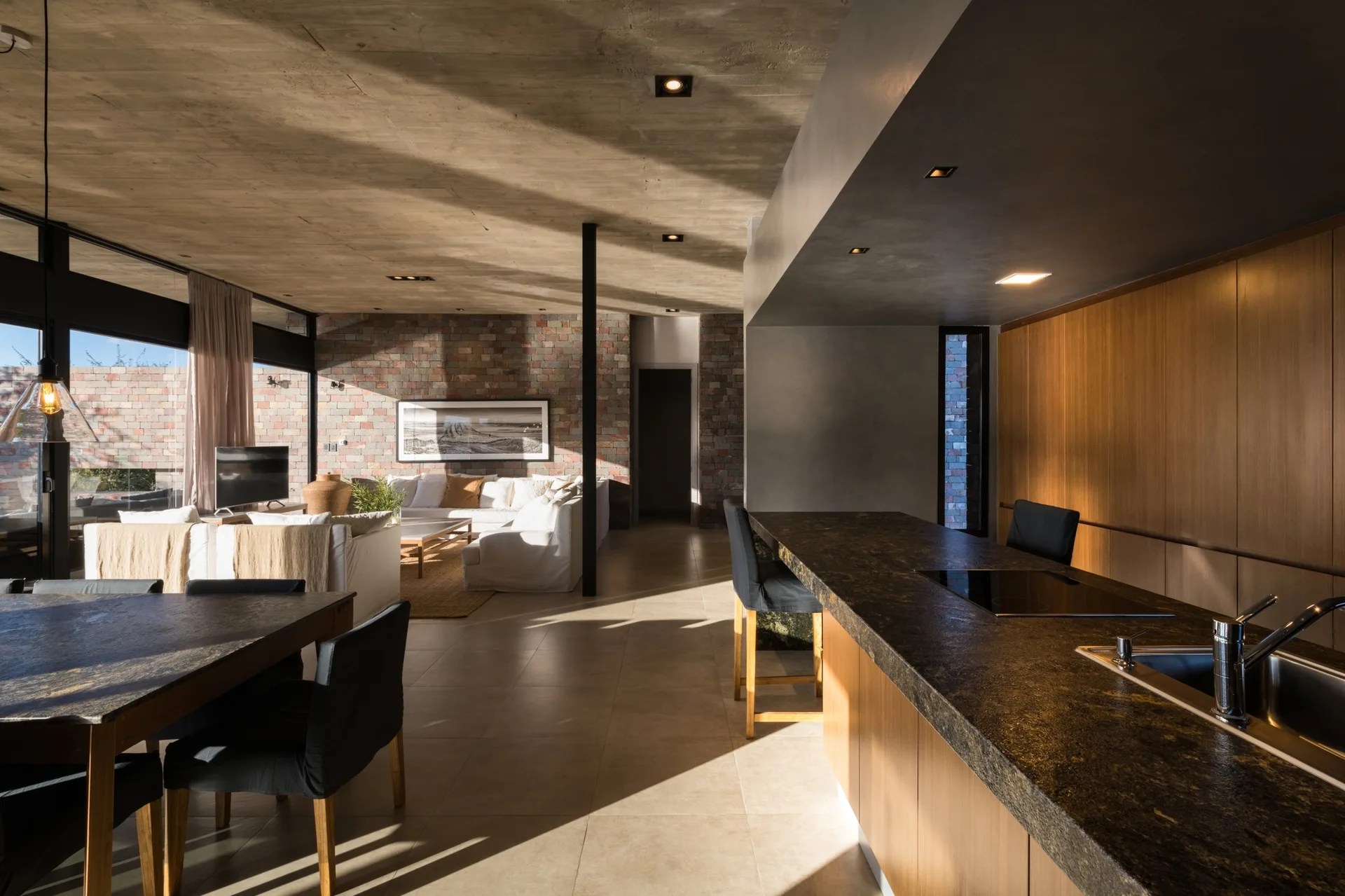 An open-plan interior featuring a dark stone kitchen island in the foreground and a living area with an exposed brick wall in the background.