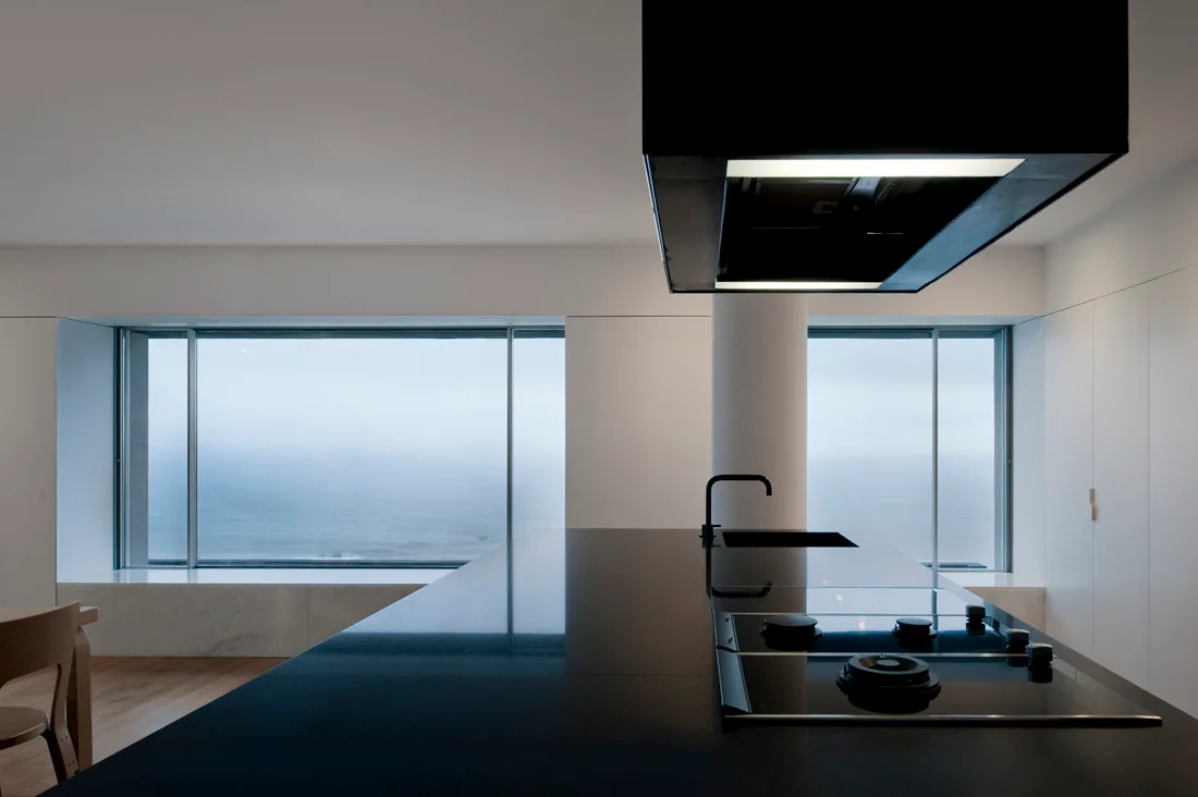 A modern kitchen featuring a sleek black island and range hood set against white walls and large windows overlooking a misty ocean view.