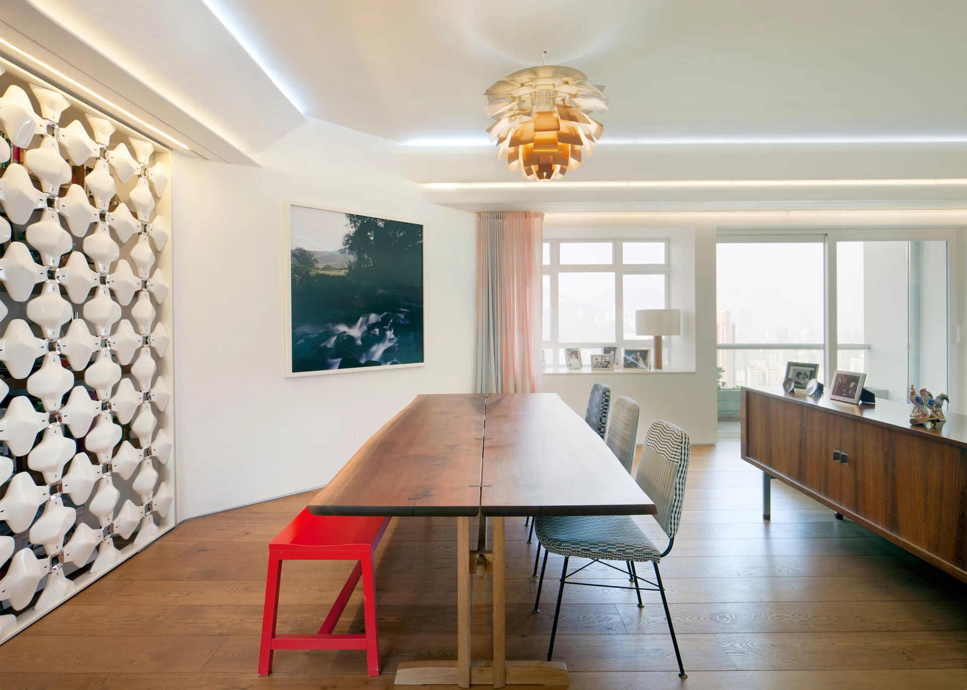 A bright dining room features a long live-edge wood table, a red bench, patterned chairs, and a white geometric wall screen.