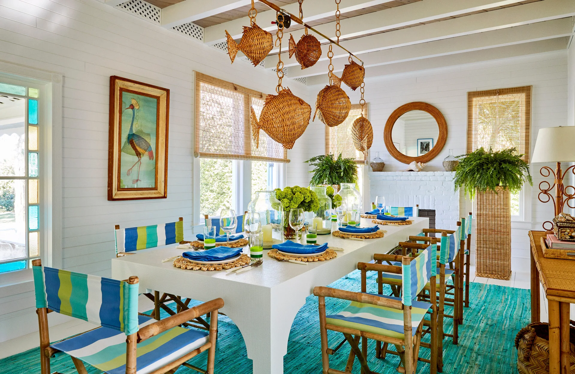 A coastal dining room features a white table surrounded by striped director chairs under fish-shaped wicker pendant lights.