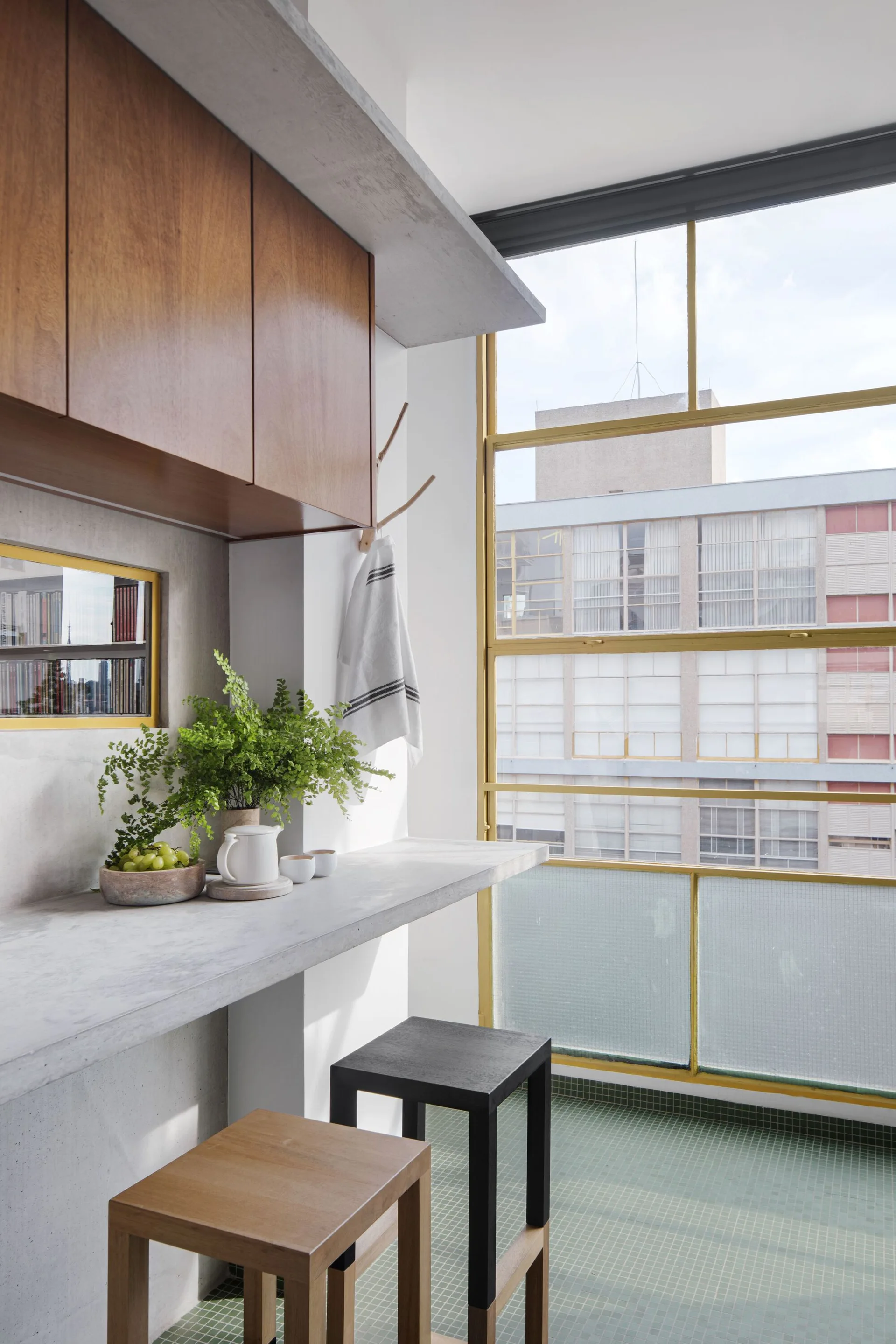 A concrete breakfast bar with two stools stands next to a large window with yellow frames and green tiled flooring.