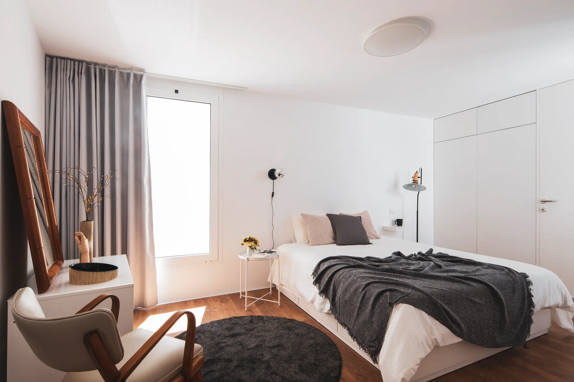 A minimalist bedroom features a bed with grey bedding, a built-in white wardrobe, a wood-framed floor mirror, and a mid-century armchair.