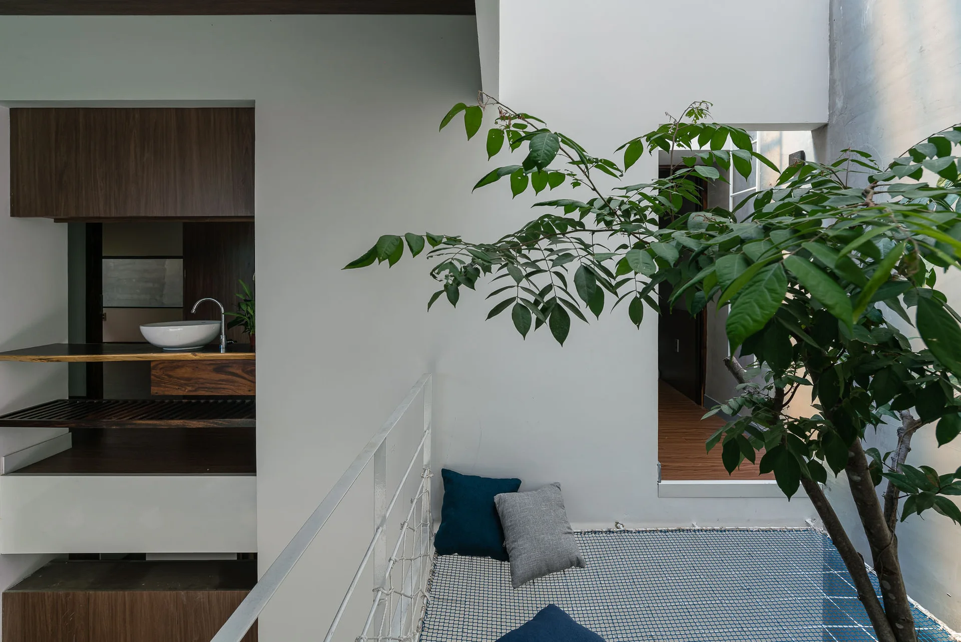 A minimalist white loft landing features a structural netting floor hammock with blue pillows next to an indoor tree and a wooden vanity sink niche.