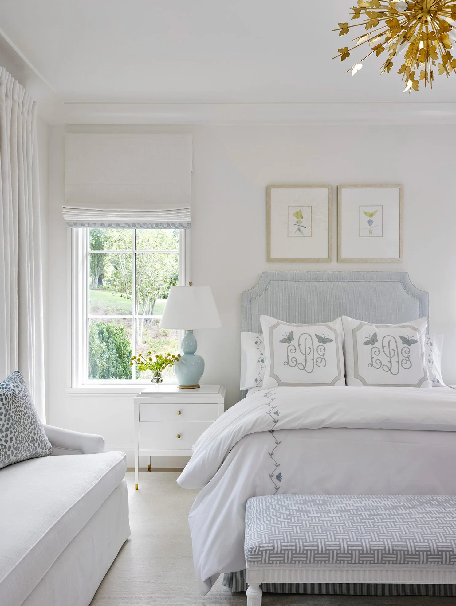 A bright white bedroom features a light blue upholstered bed with monogrammed pillows, a matching table lamp, and a patterned bench at the foot of the bed.