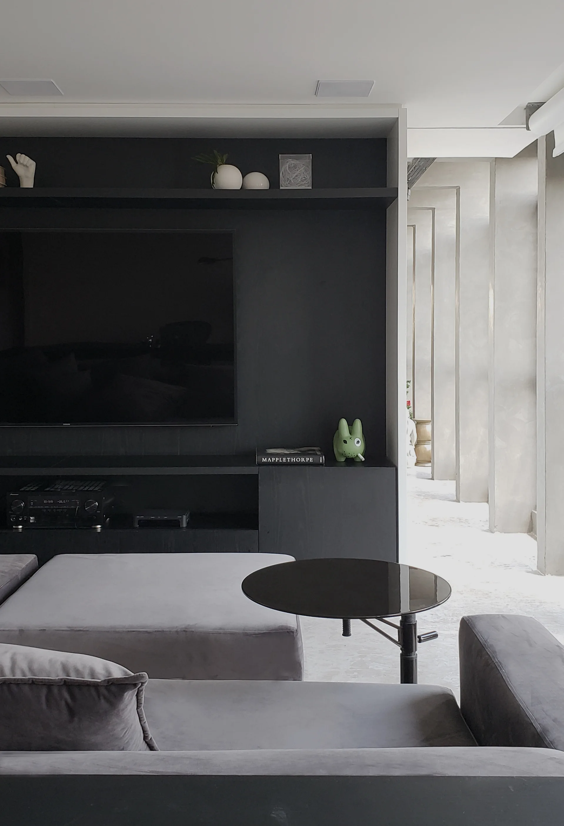 A modern living room features a grey sectional sofa, a round side table, and a built-in matte black media cabinet with a wall-mounted television.