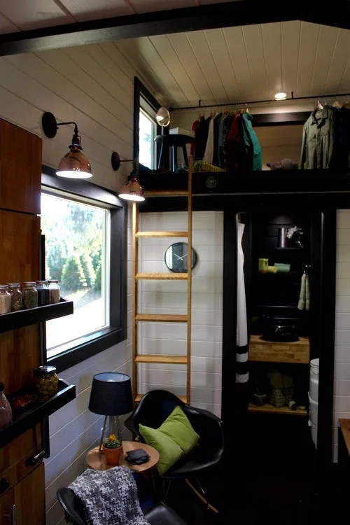 A compact living area features horizontal white plank walls, a wooden ladder leading to a sleeping loft, black trim, and a black armchair.