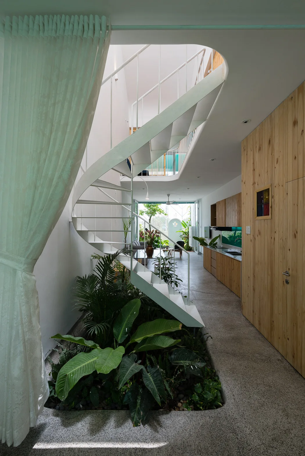 A white spiral staircase stands above an indoor garden planter set into a grey speckled floor in a modern open-plan home.