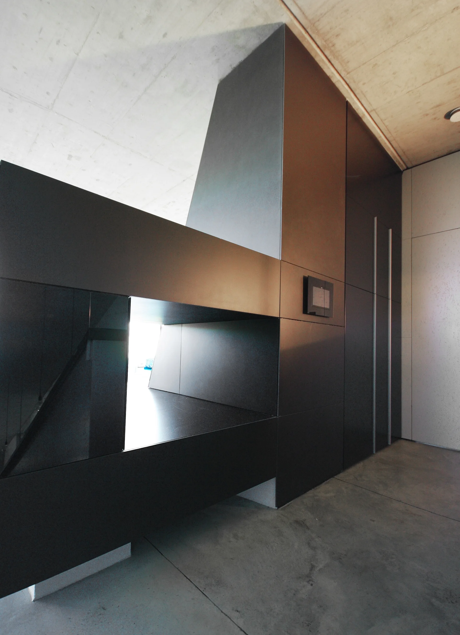 A large dark gray minimalist cabinetry unit with a recessed niche is surrounded by exposed concrete flooring and ceiling.