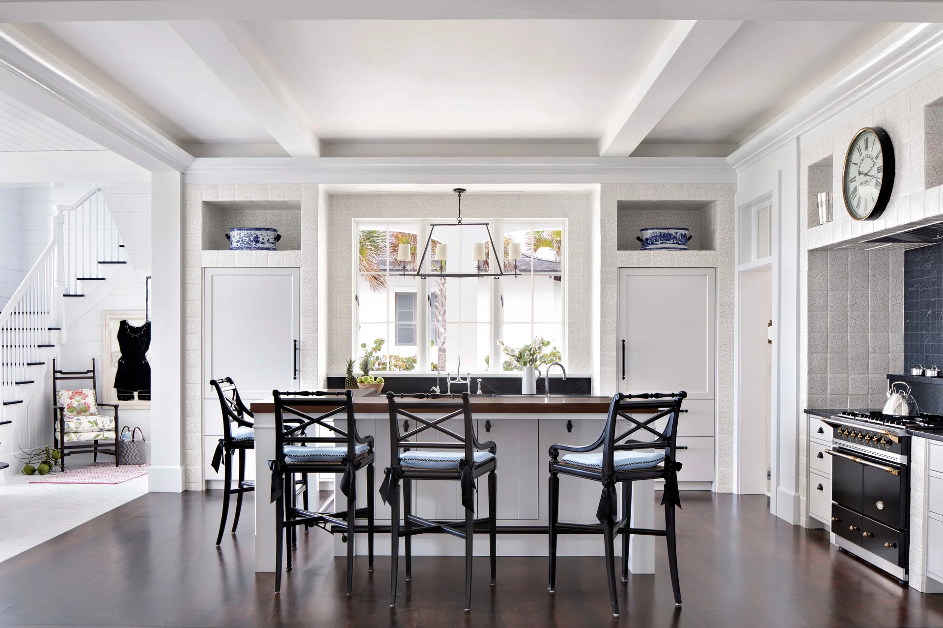 A bright kitchen features a white central island with black x-back stools, white cabinetry, dark wood flooring, and a large window.