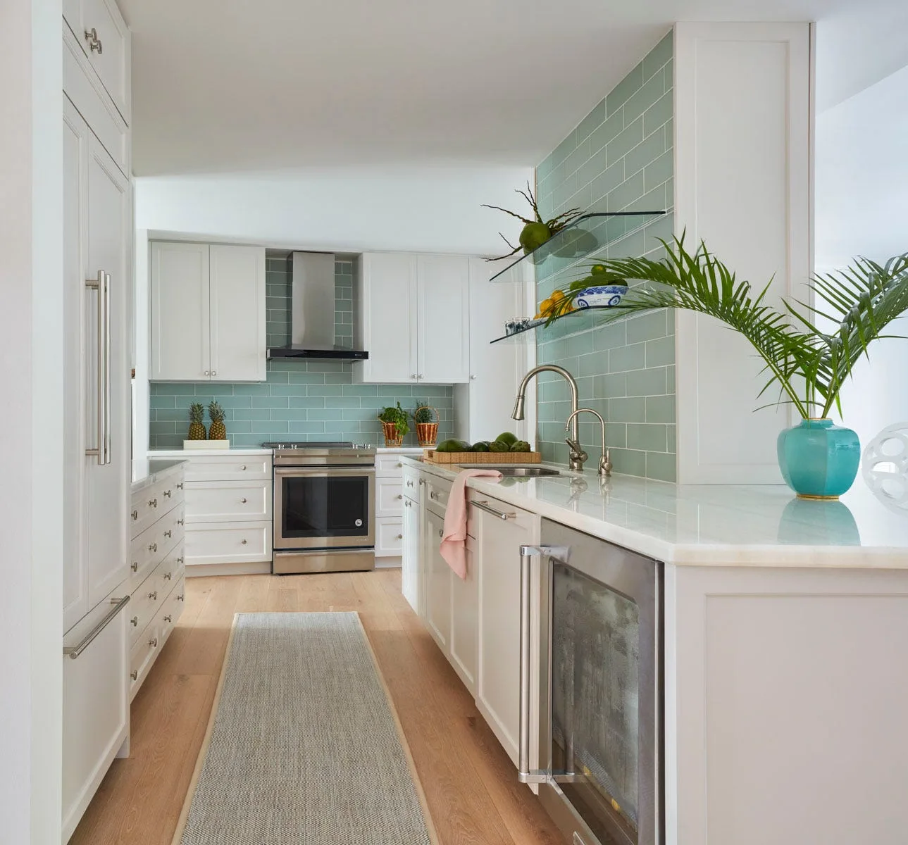 A galley kitchen features white shaker cabinets, a light green subway tile backsplash, glass floating shelves, and light wood floors with a long runner.