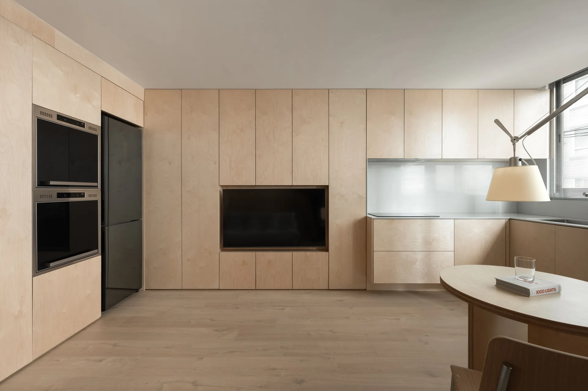 A minimalist kitchen features seamless light wood cabinetry, a recessed television, integrated black appliances, and a circular dining table.