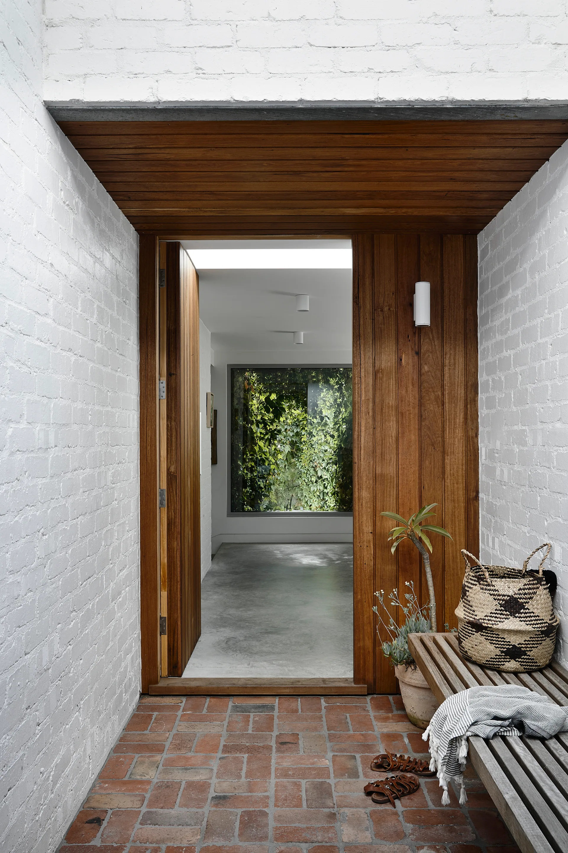 A transitional entryway featuring white brick walls, a wood slatted bench, and a view into an interior hallway with a large window covered in green ivy.