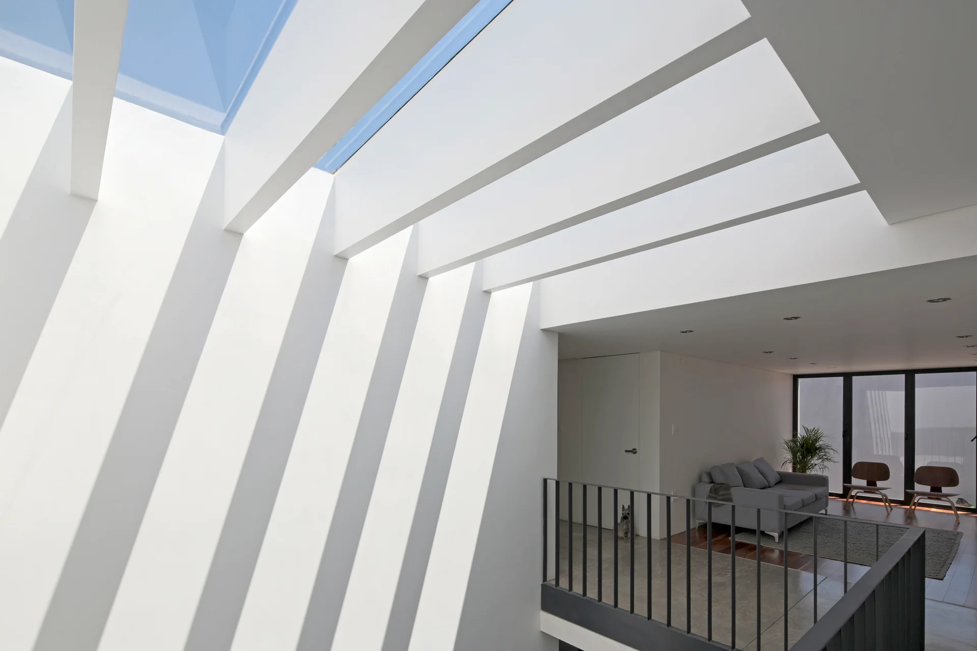 Large white ceiling beams under a skylight cast linear shadows onto a white wall above a modern mezzanine living room with a grey sofa.