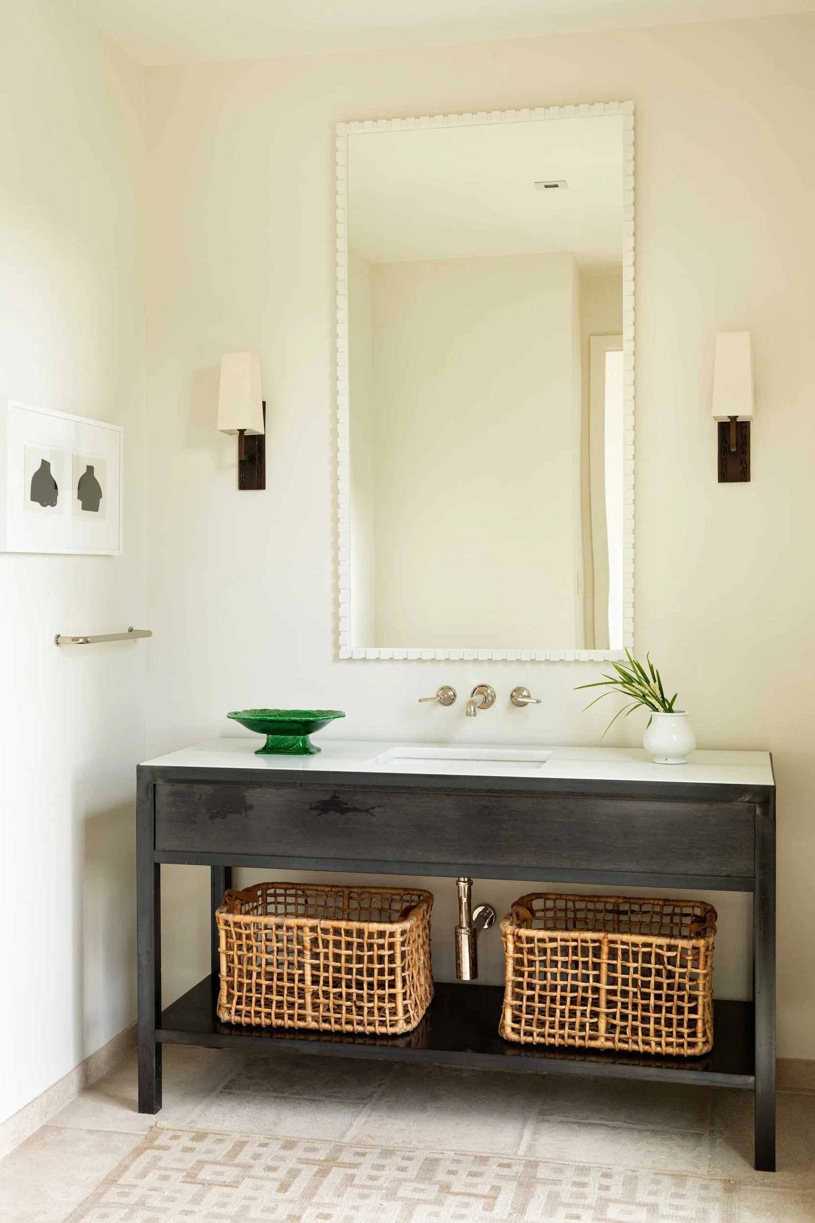 A powder room features a black console vanity with a marble countertop, wall-mounted faucet, a white-framed rectangular mirror, and two wicker baskets below.