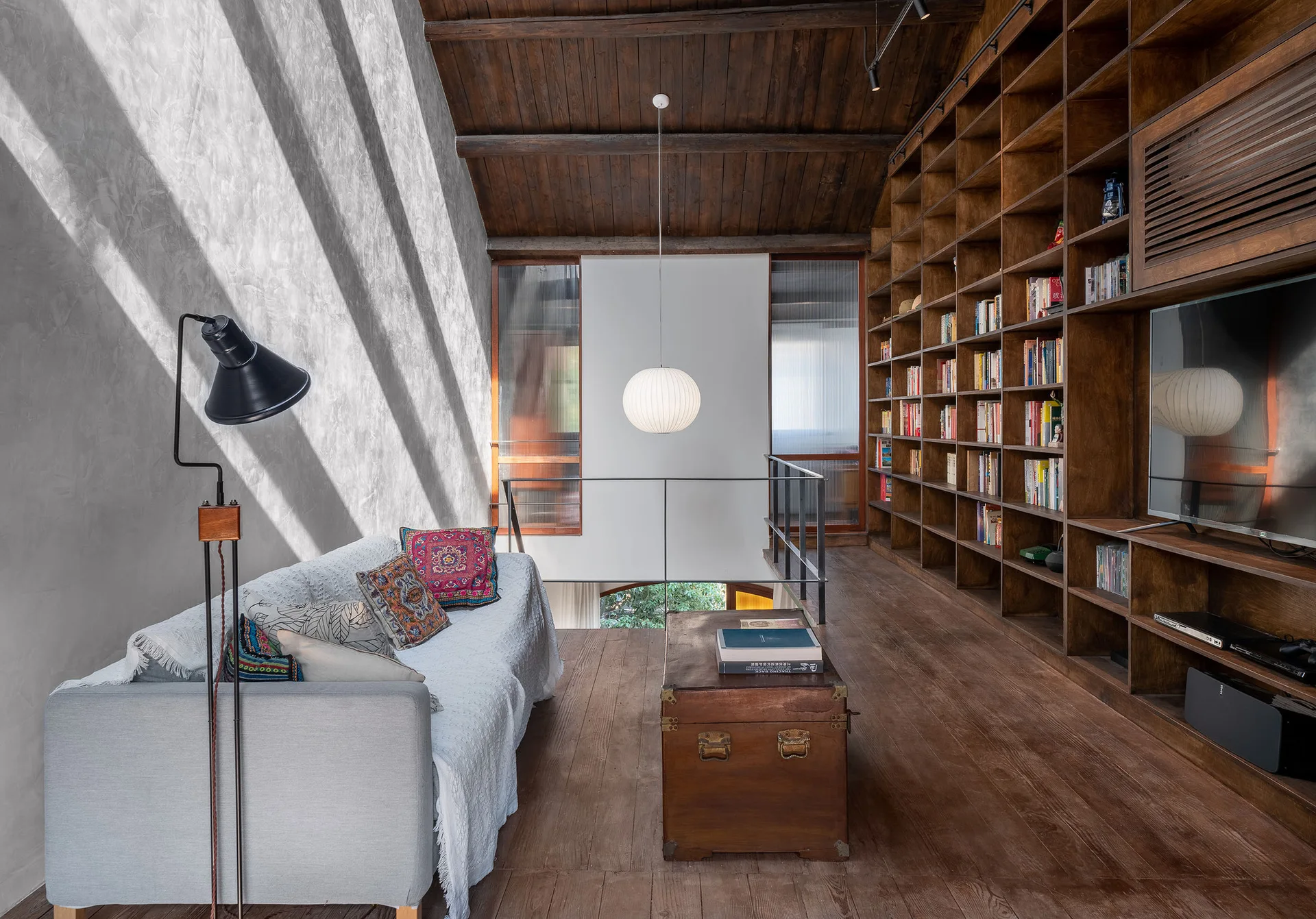 A loft living room features a grey sofa under a sloped wood ceiling, a vintage trunk table, and a massive dark wood built-in bookcase.