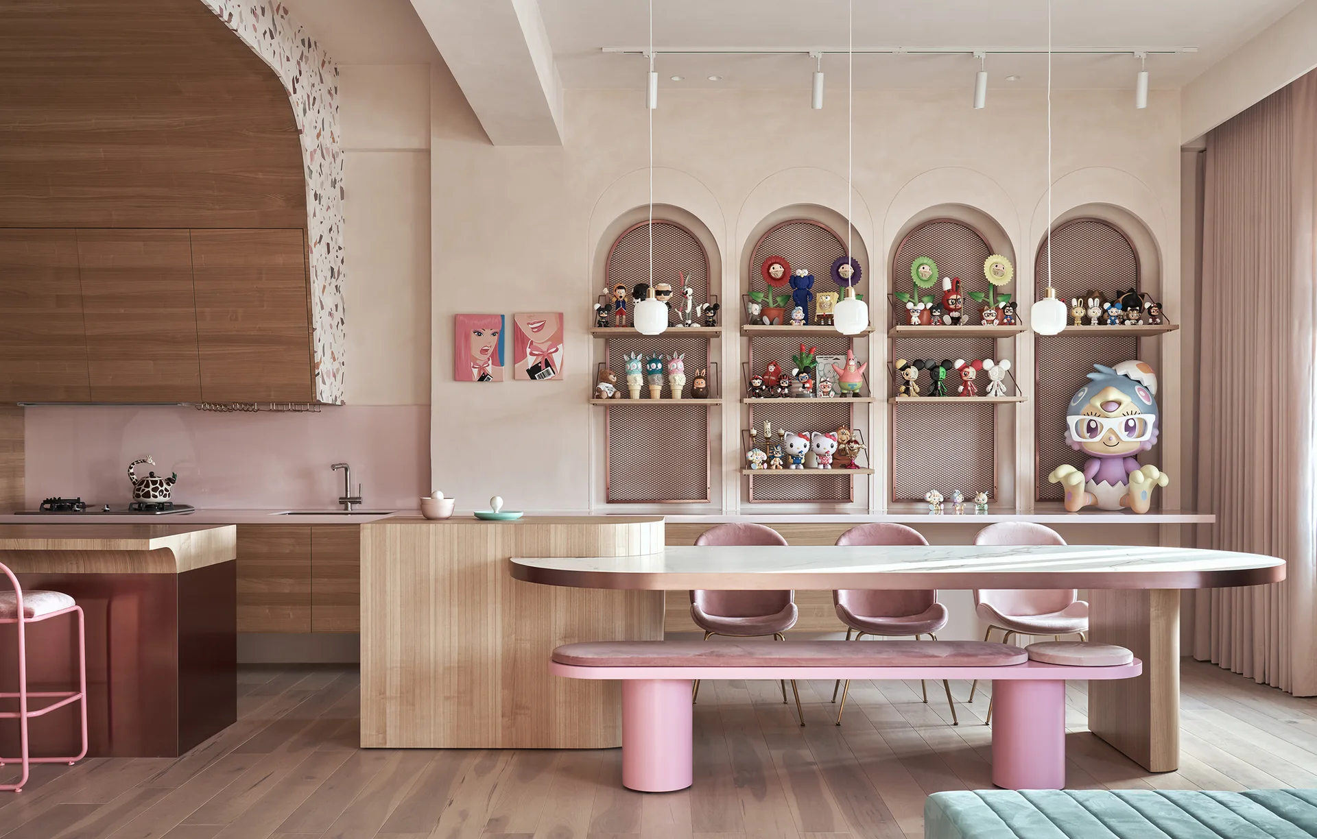 A blush pink kitchen and dining area features an integrated wood island table, pink velvet seating, and arched wall niches with collectible figurines.
