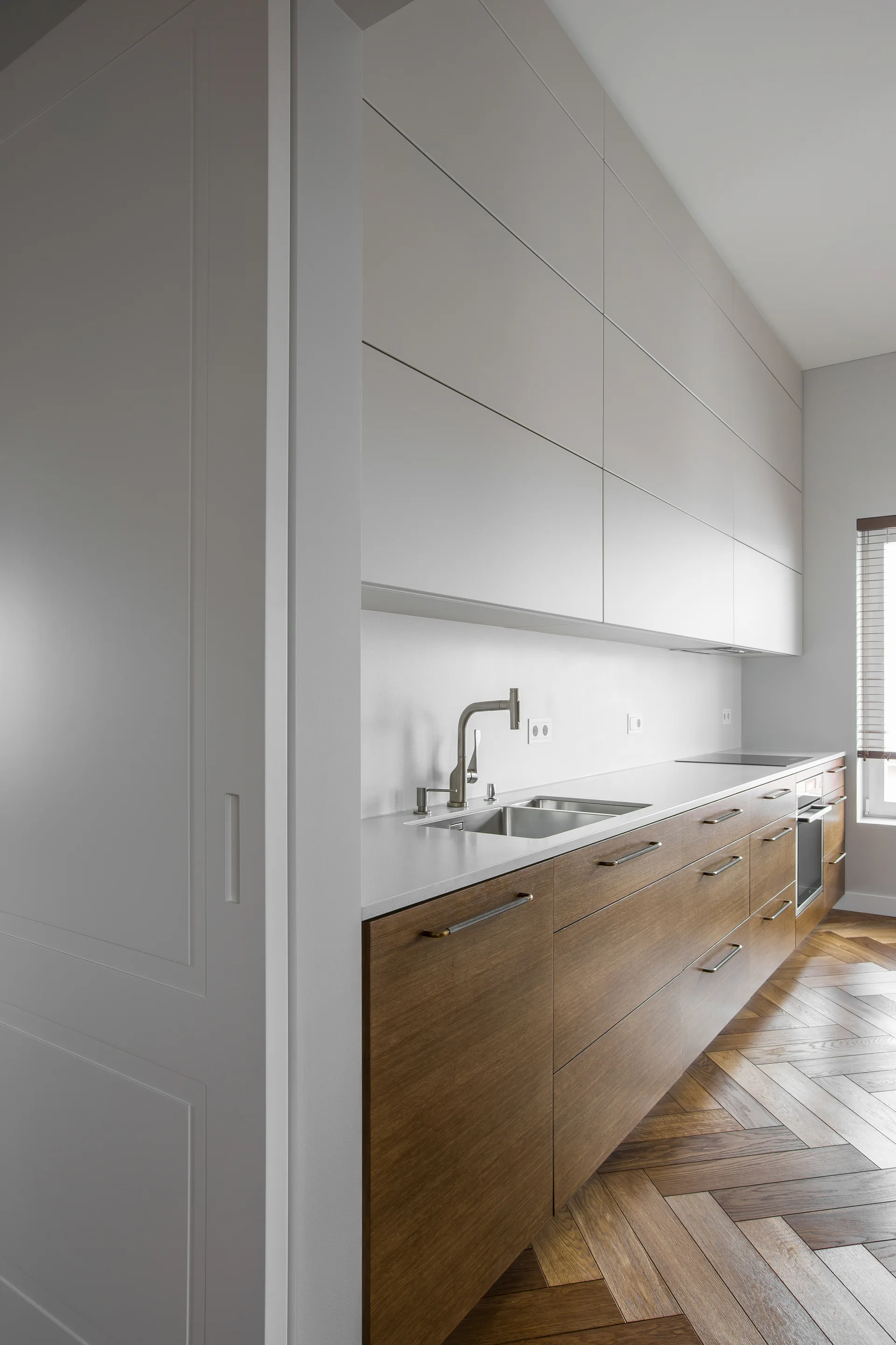 A minimalist kitchen featuring two-tone cabinets with wood base drawers, matte off-white upper cabinets, and a herringbone hardwood floor.