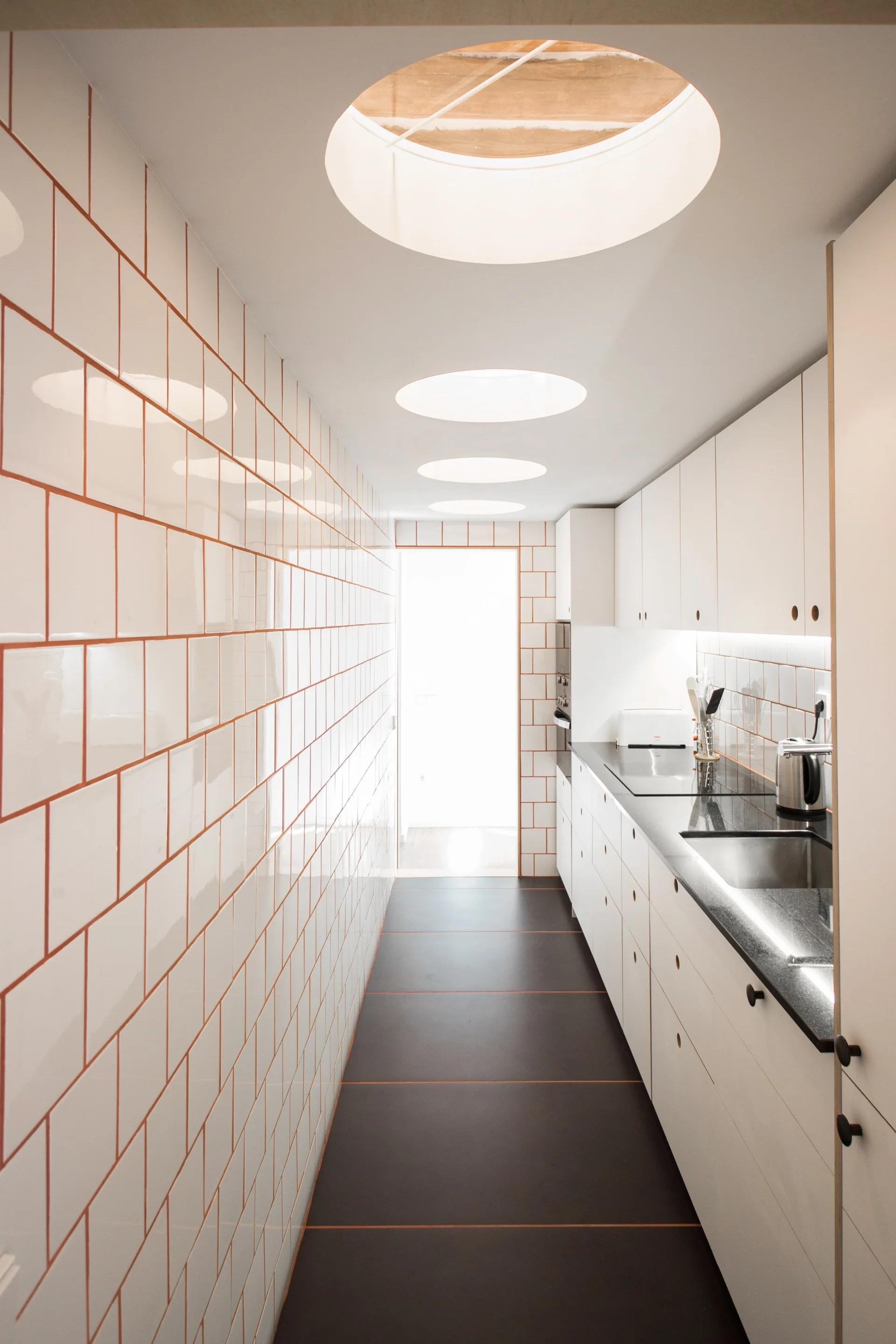 A galley kitchen featuring a wall of white square tiles with warm grout, white cabinets, dark countertops, and three circular skylights.