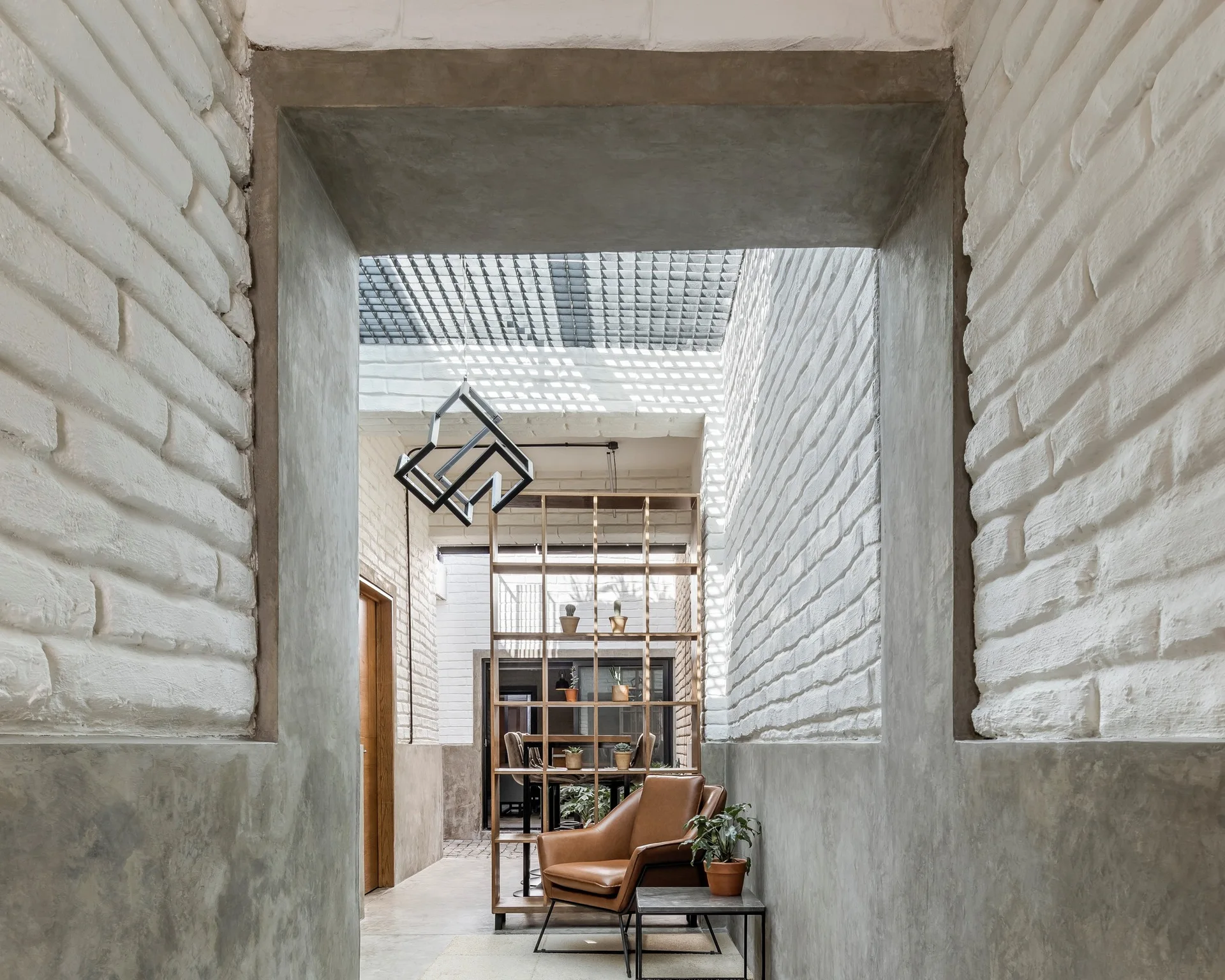 An interior view framed by a concrete doorway shows a brown leather armchair next to a tall wooden shelving unit against white painted brick walls.