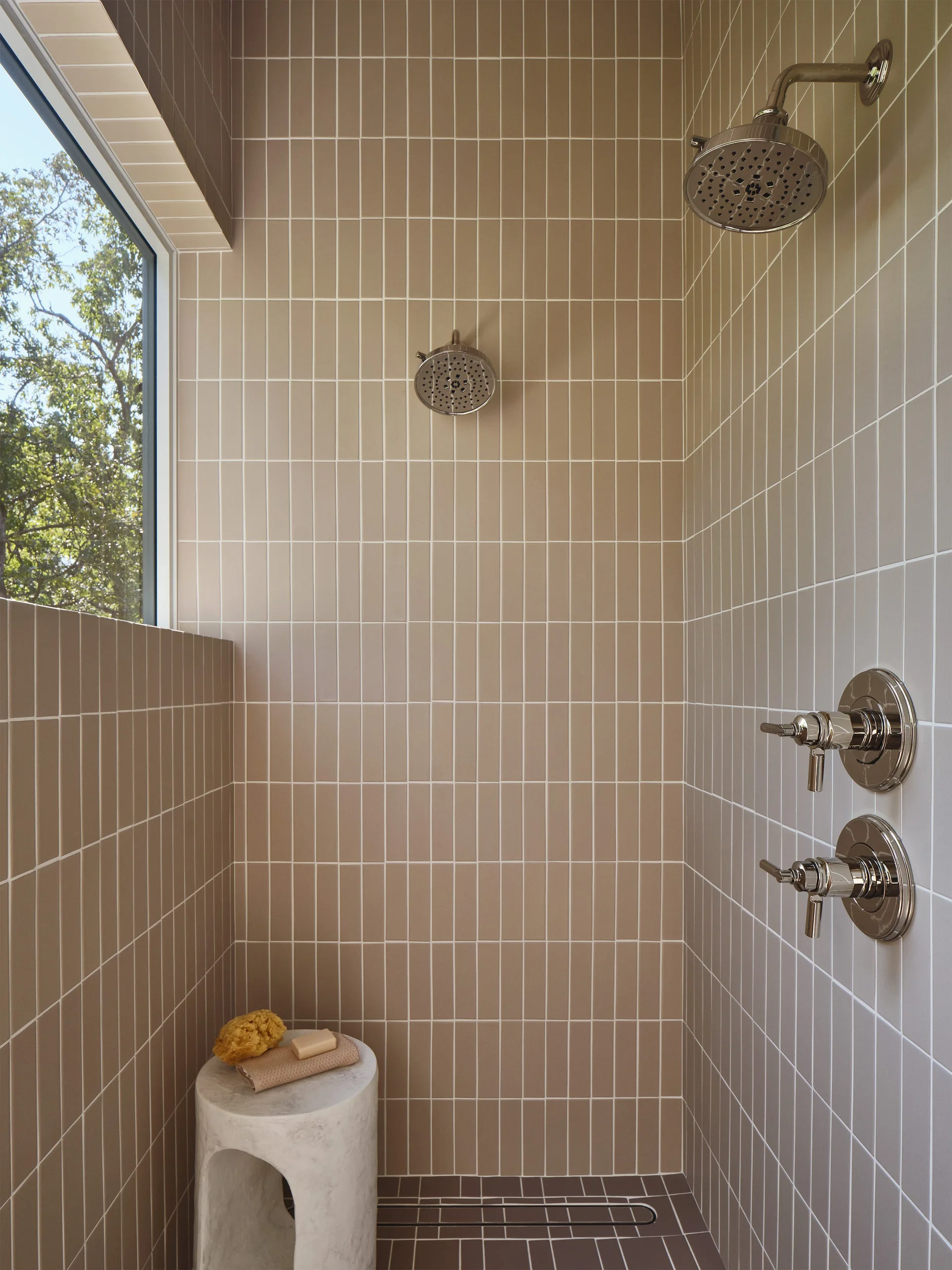 A modern shower with vertical beige wall tiles features dual chrome shower heads, a linear floor drain, and a small stone stool.