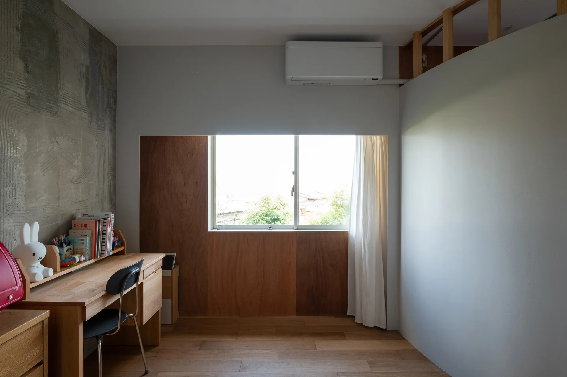 A minimalist kids study space features a wooden desk with a bunny lamp, a textured concrete accent wall, and a wood-paneled recessed window.
