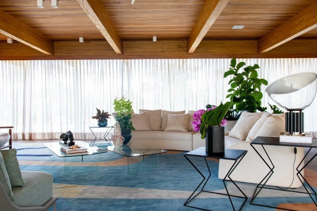A cream sectional sofa sits on a large blue patterned rug beneath an exposed wood beam ceiling, with sheer curtains lining the background.