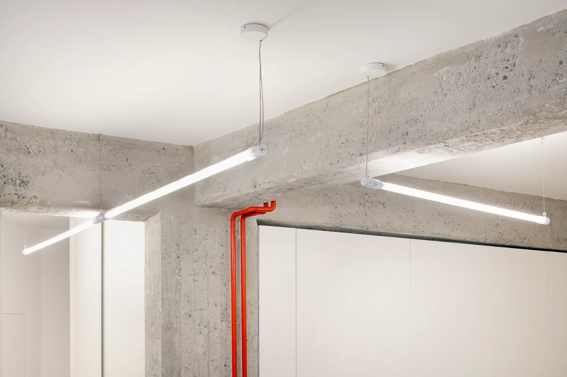 Suspended linear tube lights hang below exposed concrete ceiling beams next to a bright red vertical pipe against a white wall.