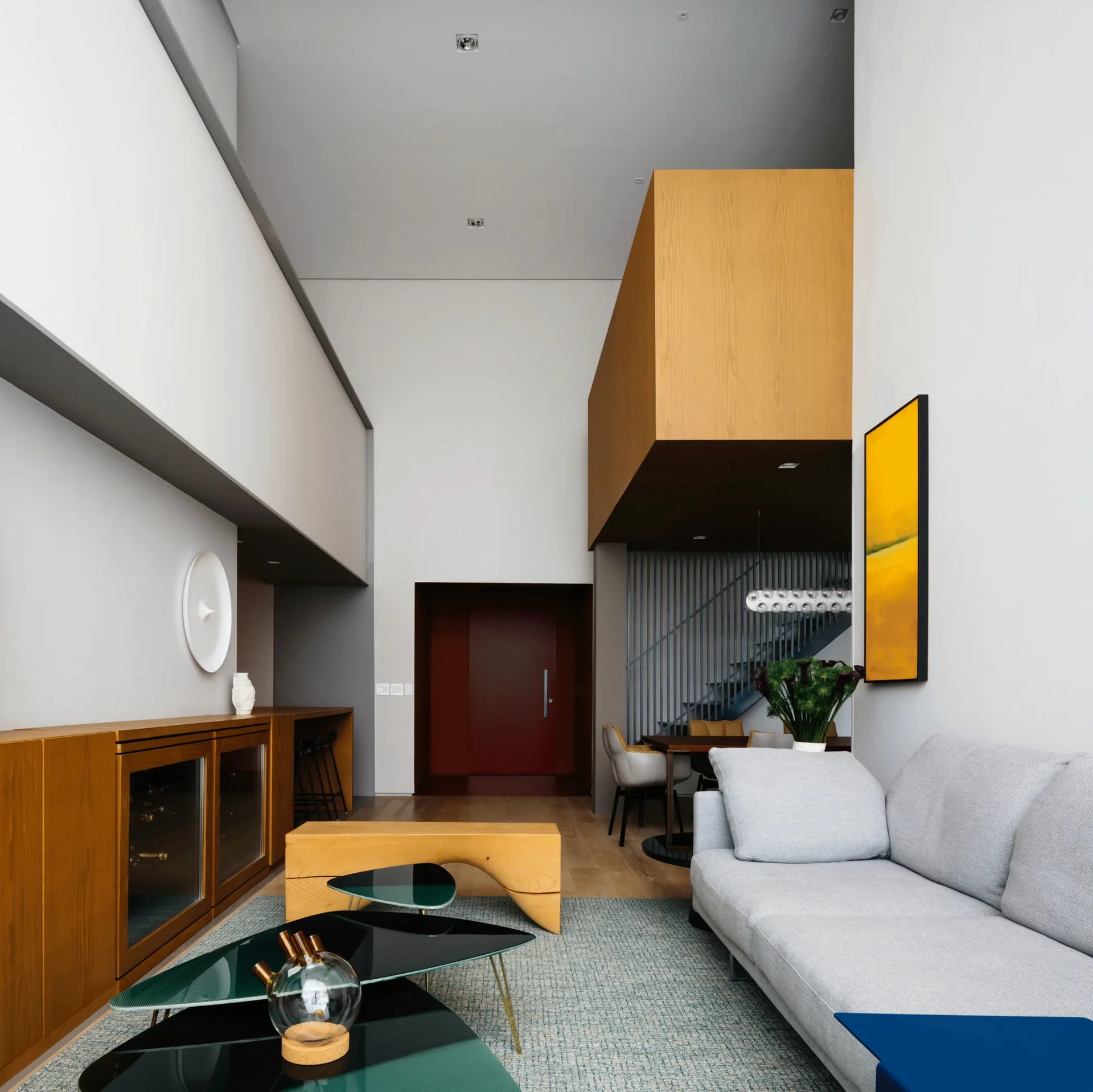 A double-height modern living room features a grey sofa, dark glass nesting tables, a long wood credenza, and an elevated wood mezzanine structure.