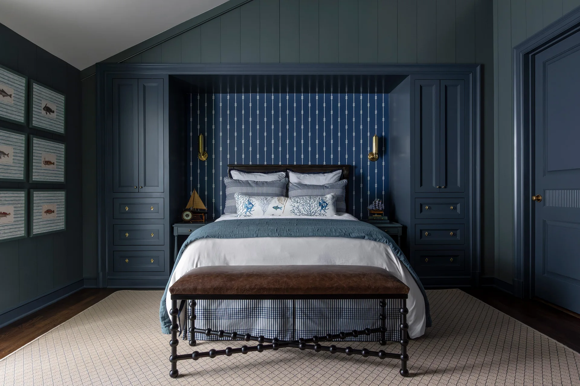 A nautical bedroom features a bed framed by custom deep blue cabinetry, vertical striped wallpaper, and a brown leather bench at the foot.