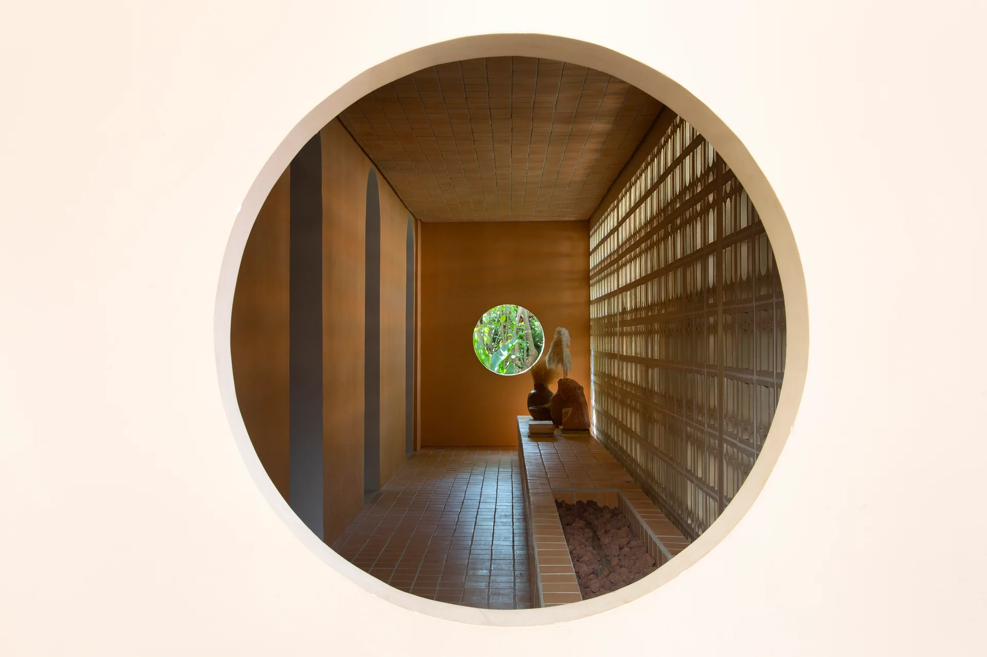 A view through a large circular wall opening looking down a terracotta tiled hallway with recessed wall arches and a patterned screen wall.