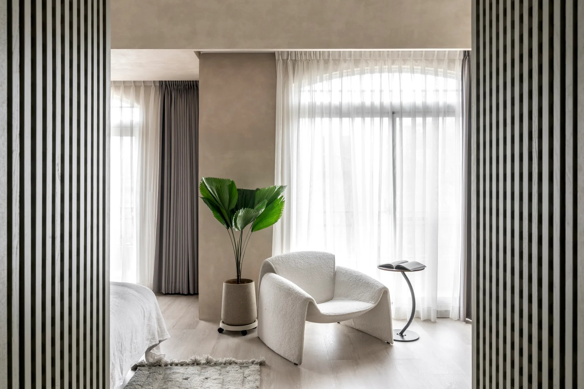 A minimalist bedroom nook with a sculptural white armchair, a curved side table, a large potted palm, and sheer curtains.