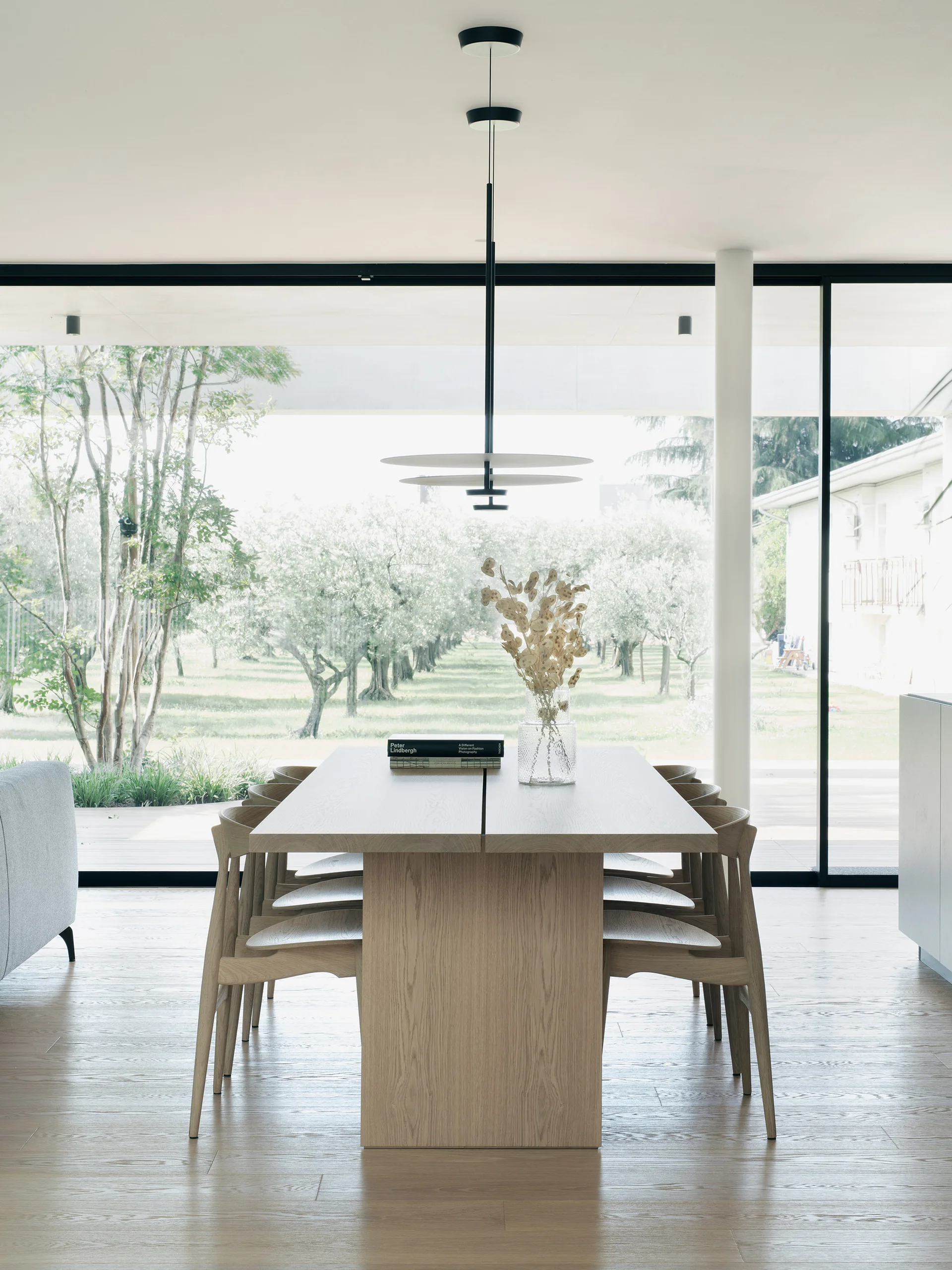 A light oak dining table with matching wooden chairs sits in front of a floor-to-ceiling glass window overlooking an outdoor tree grove.
