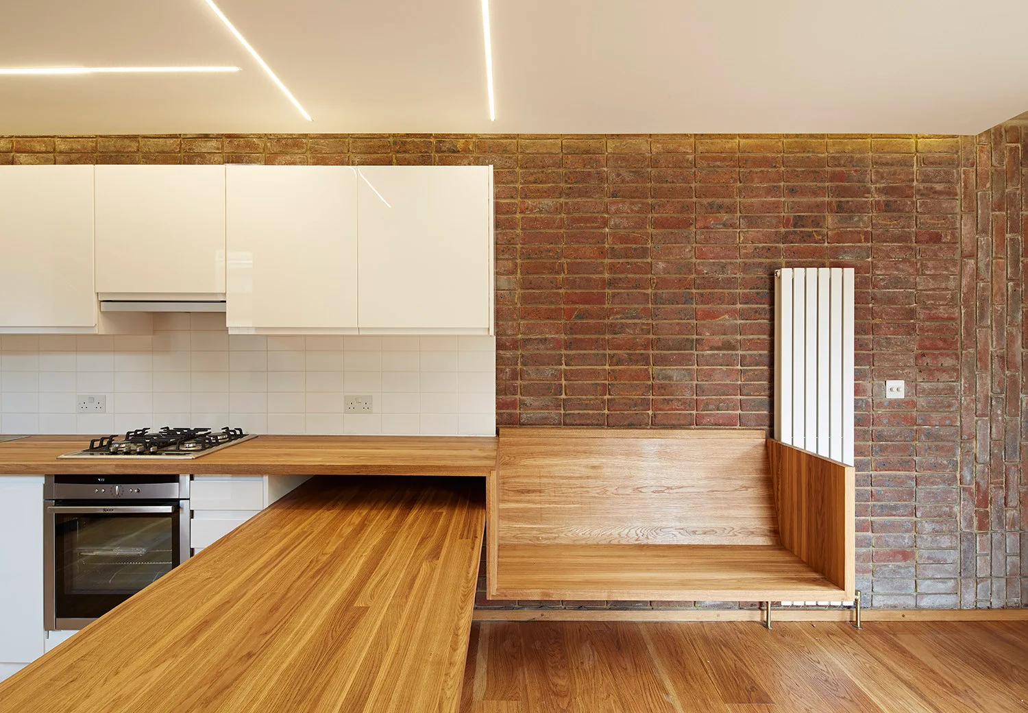 A modern kitchen featuring white upper cabinets, a wooden island countertop, a built-in wooden bench, and an exposed red brick wall.