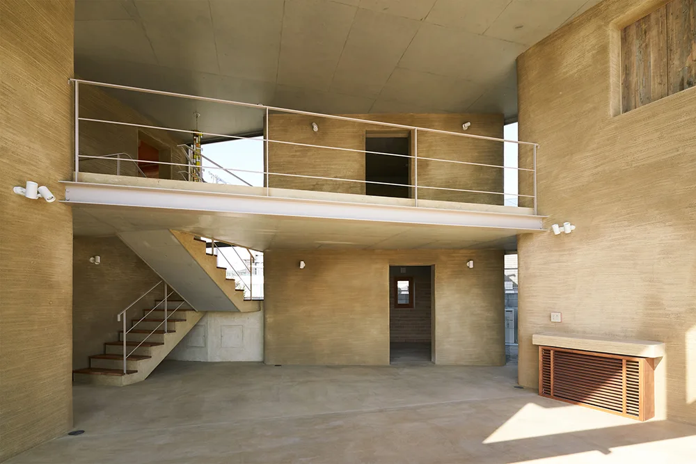 A minimalist interior showcases textured beige rammed earth walls, a steel beam mezzanine, an open staircase, and smooth grey concrete floors.