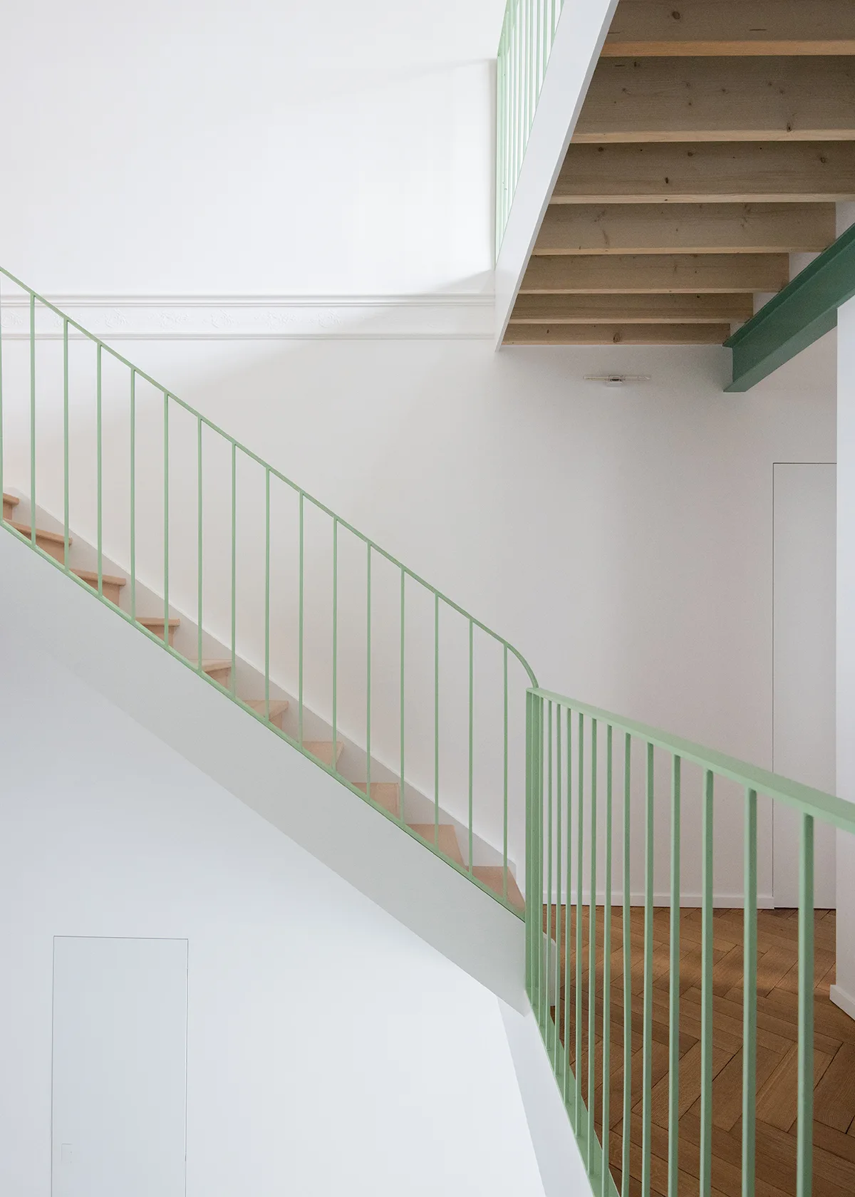 A minimalist staircase with light wood treads, a sage green spindled metal railing, and white walls showing decorative molding.