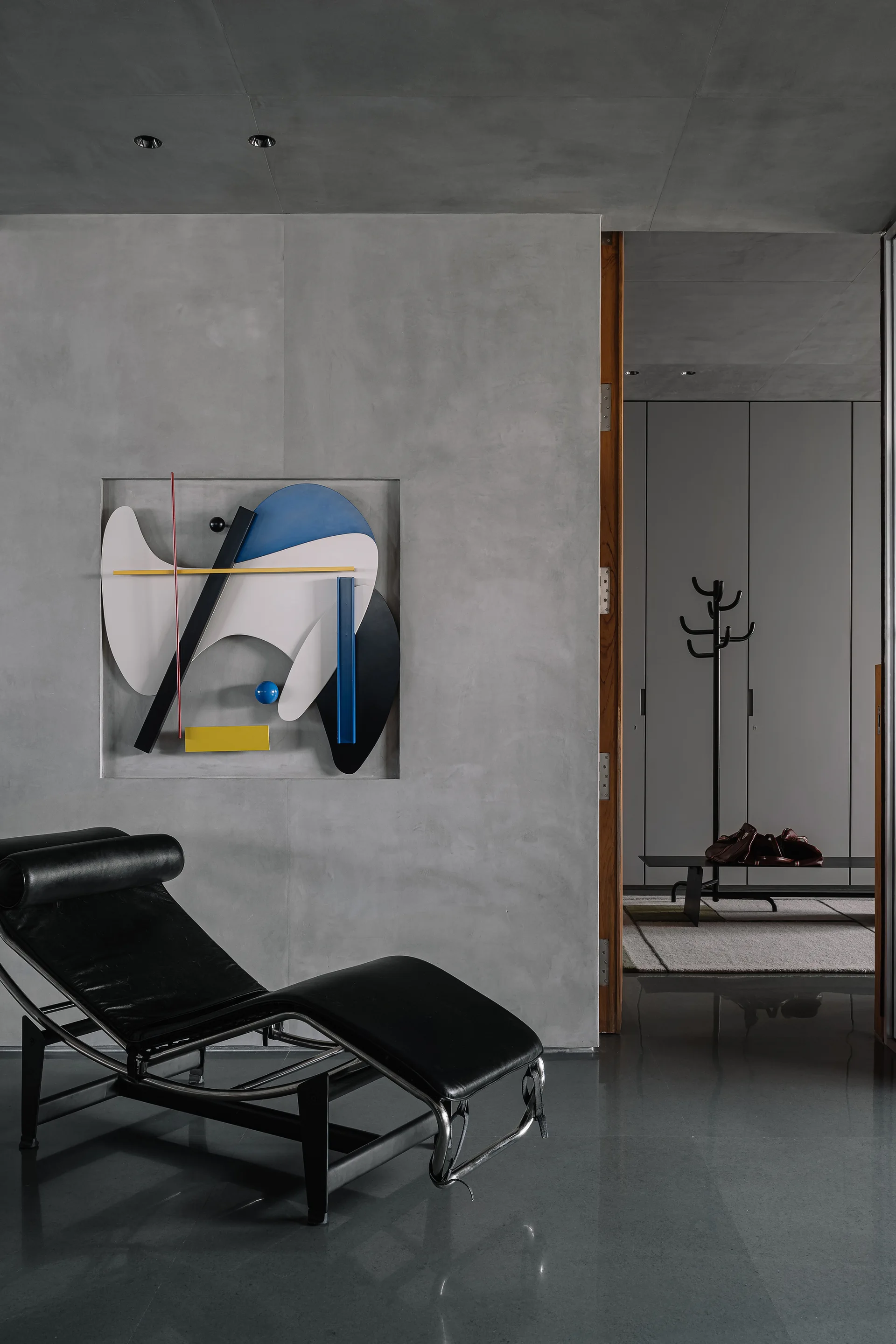 A black leather chaise longue sits on a polished floor in front of a concrete wall featuring an inset multicolored abstract geometric sculpture.