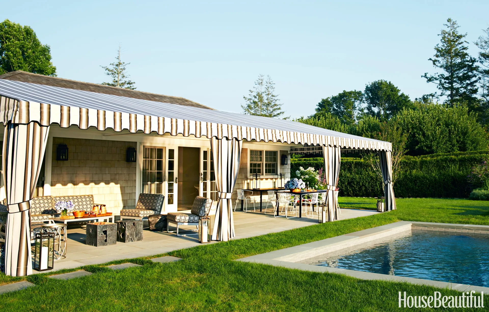 A grey stone patio features a large striped fabric awning, outdoor dining and lounge furniture, and a rectangular swimming pool surrounded by green lawn.